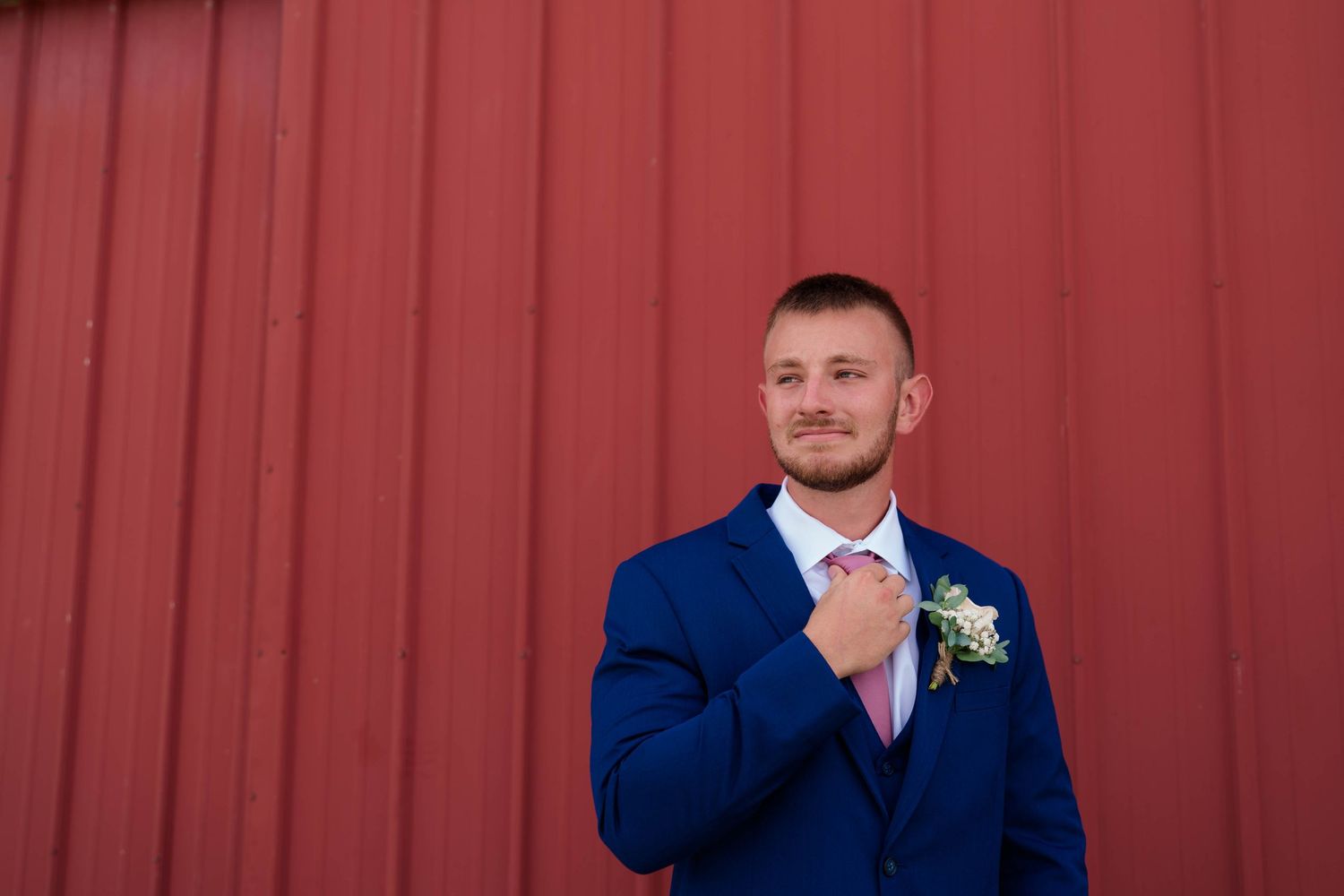 Groom in navy blue suit adjusting pink tie with boutonniere against red barn wall.