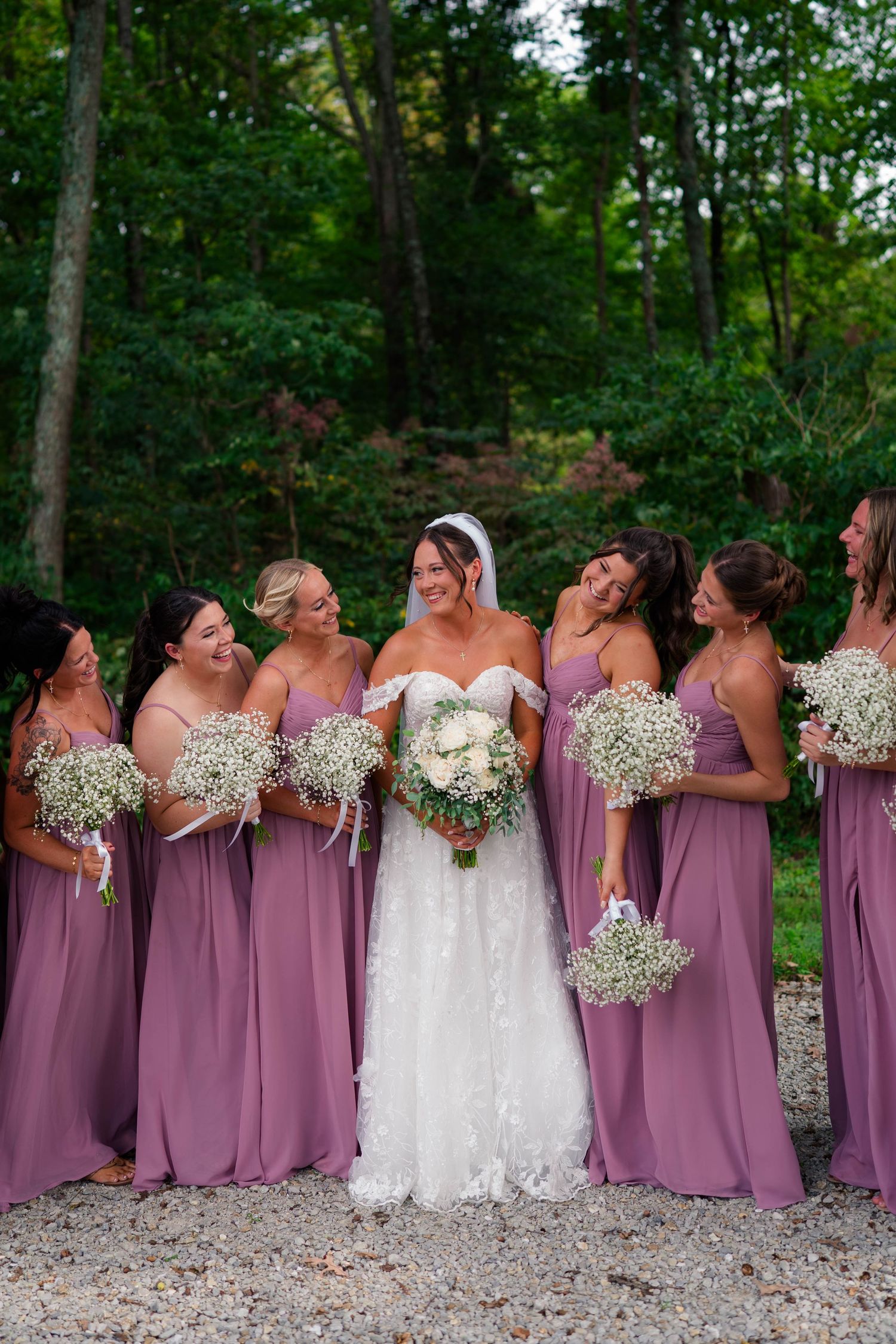 Bride in white lace gown stands with six bridesmaids in mauve dresses holding baby's breath bouquets outdoors.