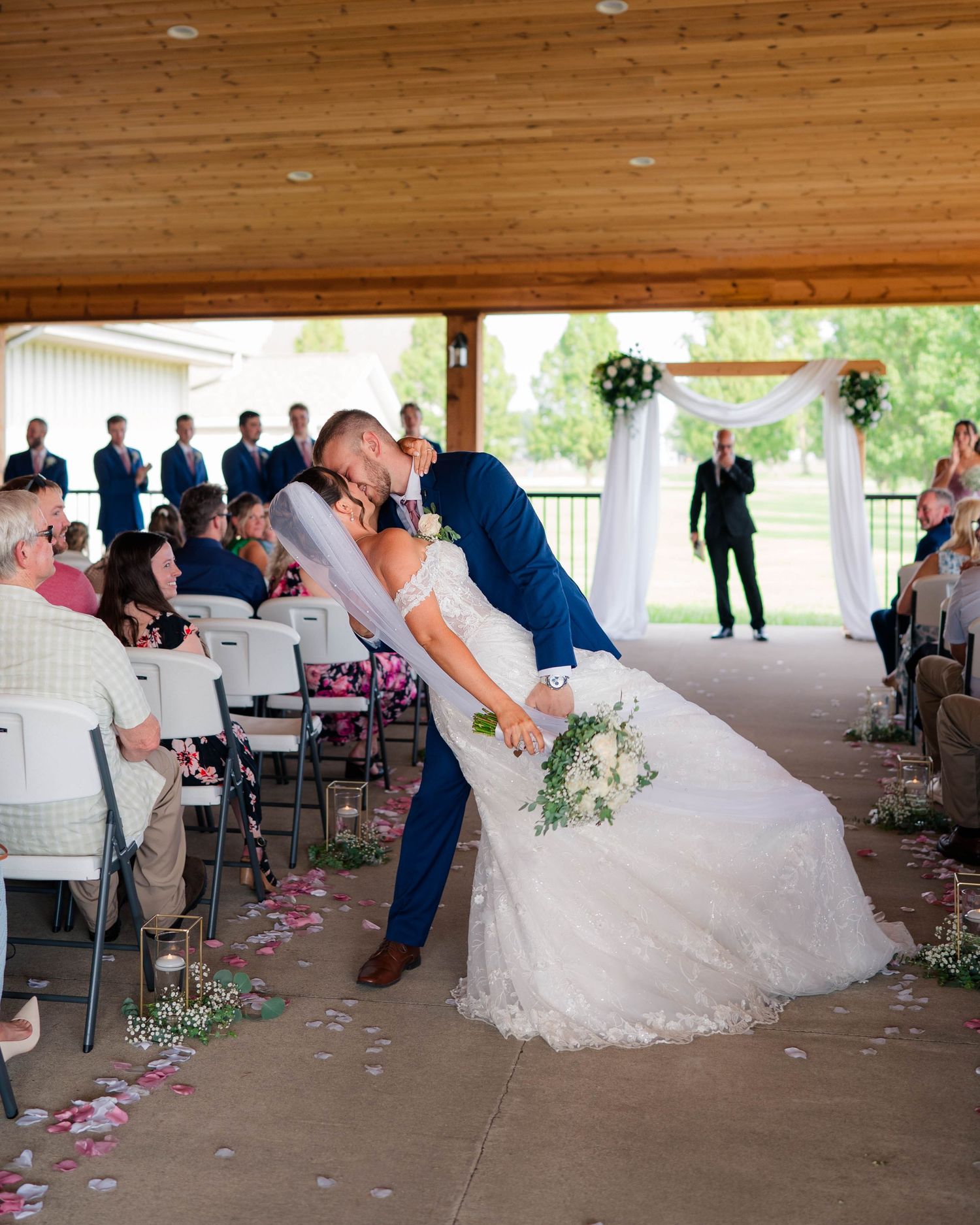 Groom dips bride for a kiss in the aisle during outdoor wedding ceremony under wooden pavilion.