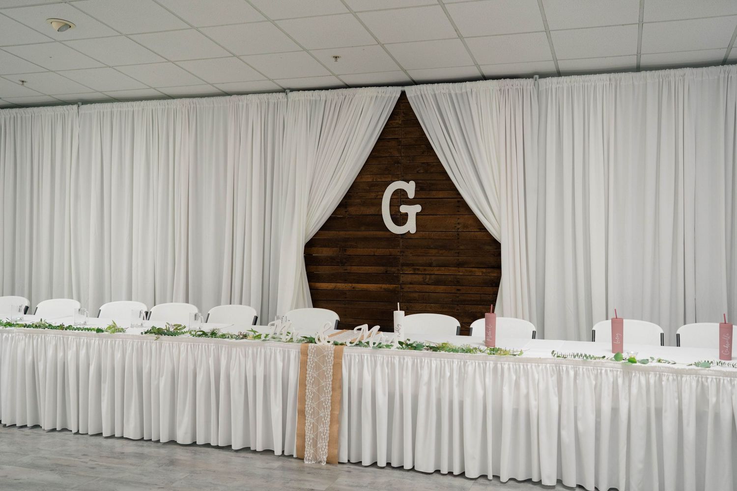 Elegant wedding reception head table with white draping and a wooden monogram letter G backdrop.