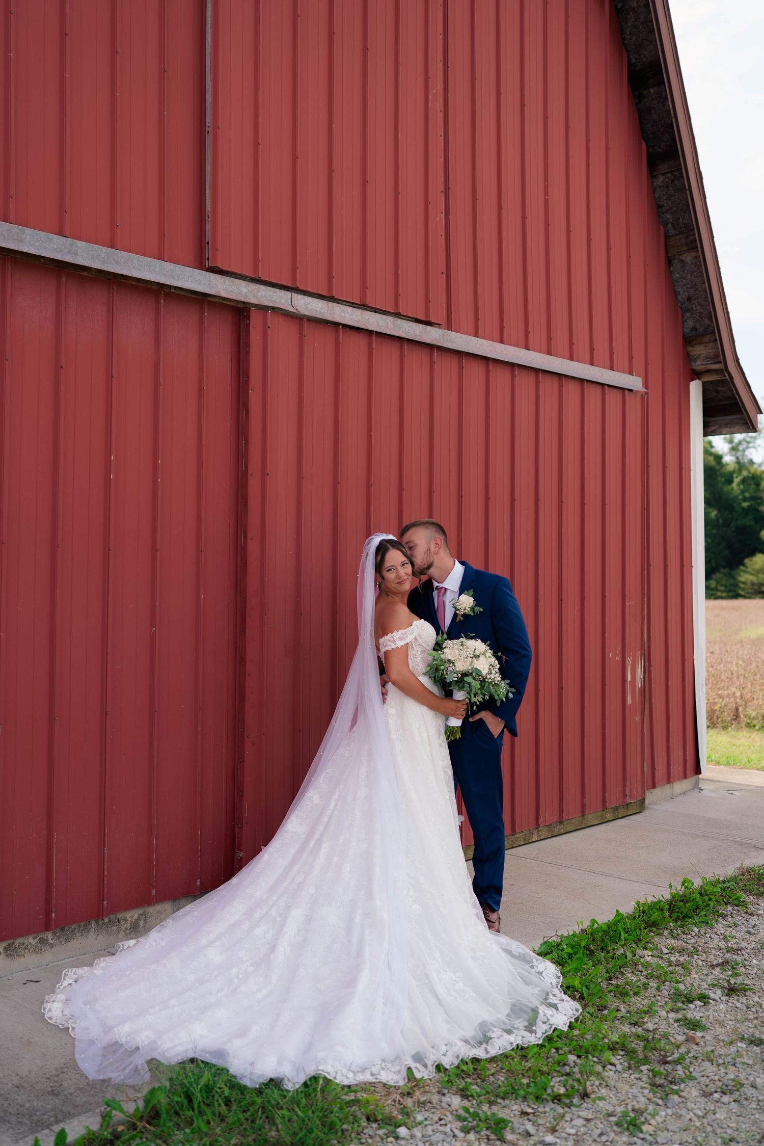 Bride and groom embrace tenderly against a red barn, her lace cathedral veil trailing on the ground.