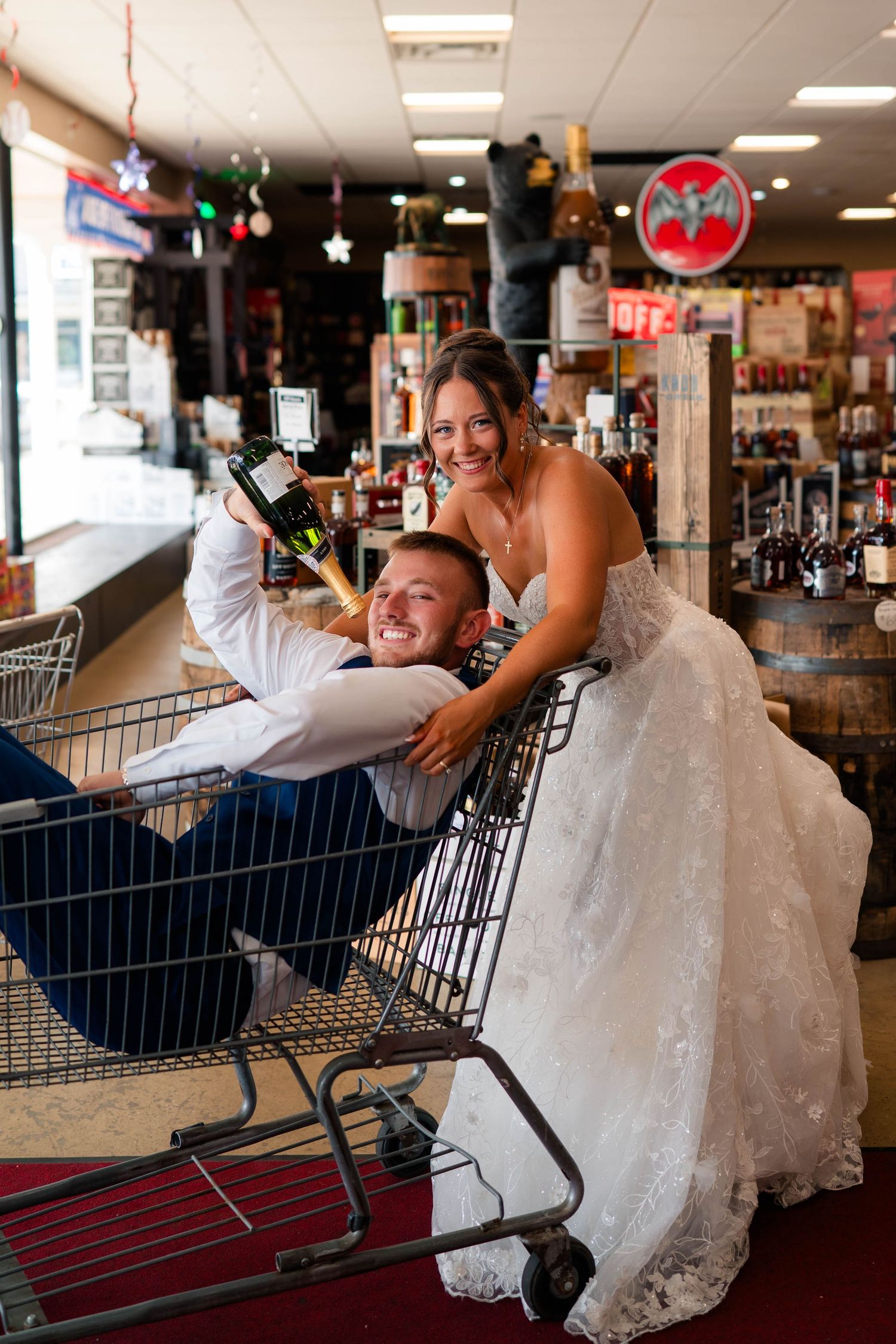 Smiling bride pushes laughing groom in shopping cart inside liquor store during fun post-wedding photo session.
