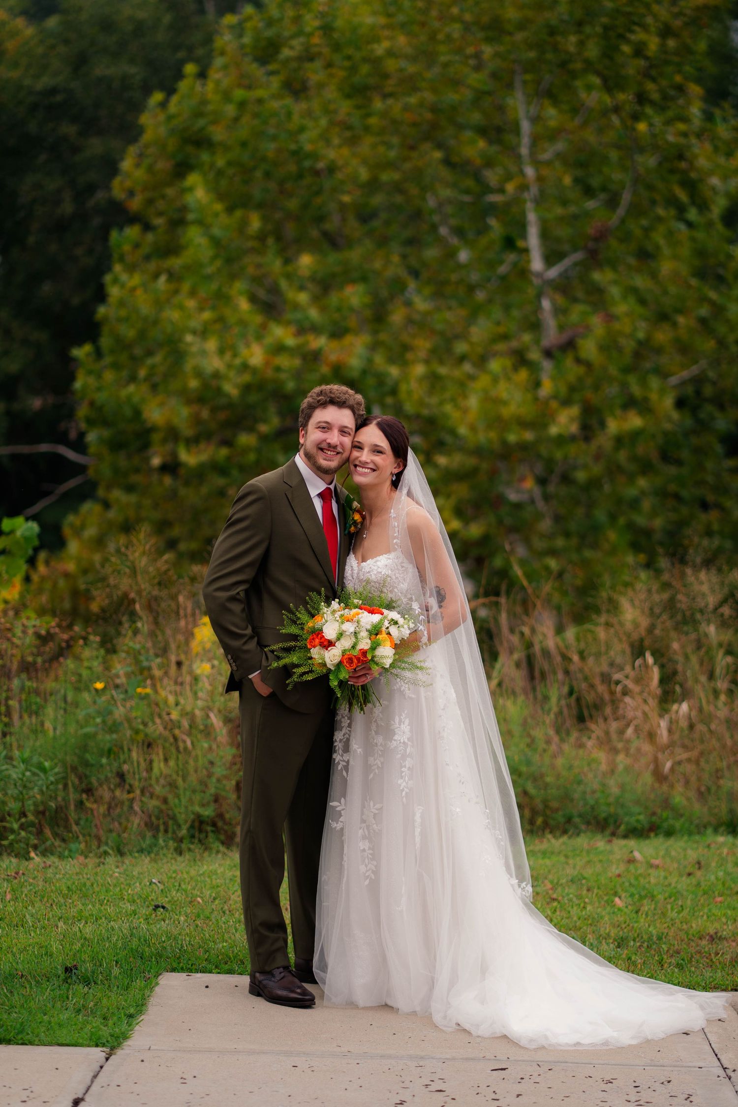 Bride and groom smiling outdoors with colorful autumn foliage and bridal bouquet.