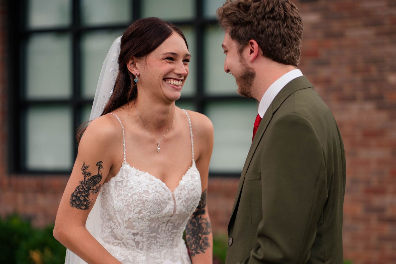 Smiling bride in lace gown with tattoos shares a joyful first look moment with groom in olive green suit.