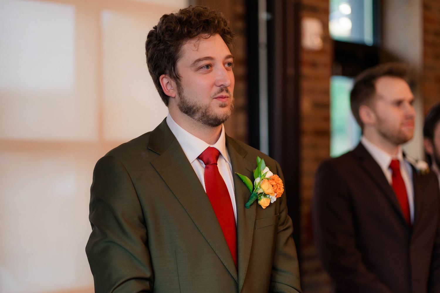 Groom in olive green suit with red tie and orange boutonniere stands at wedding altar.