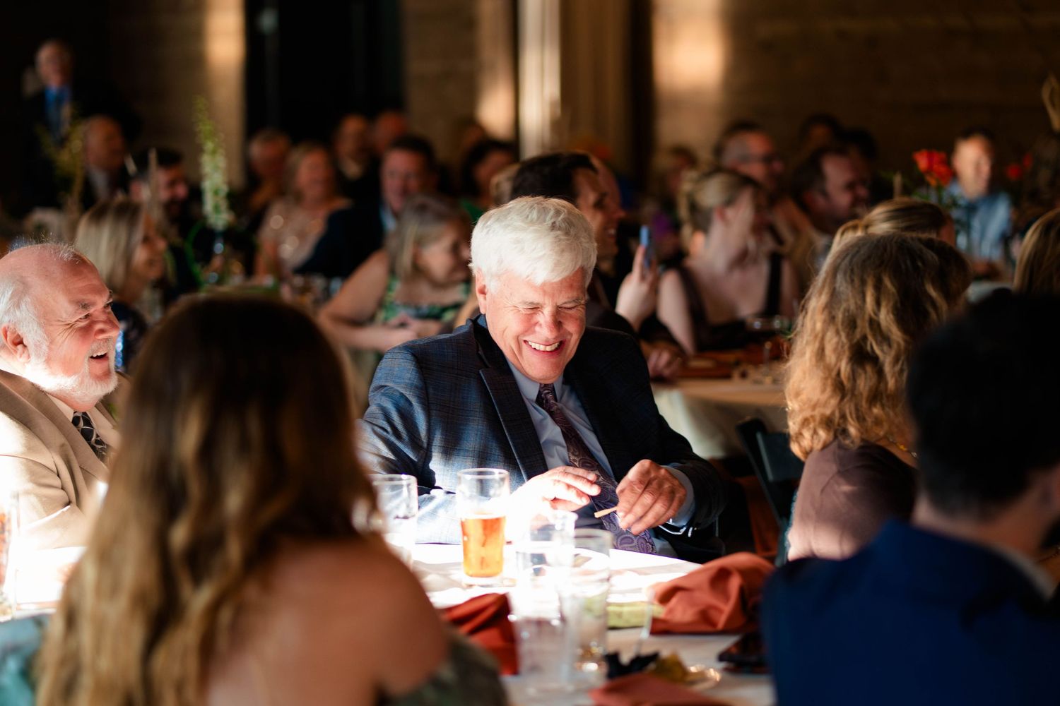 Smiling wedding guest in blue suit laughing at reception dinner table surrounded by other guests.