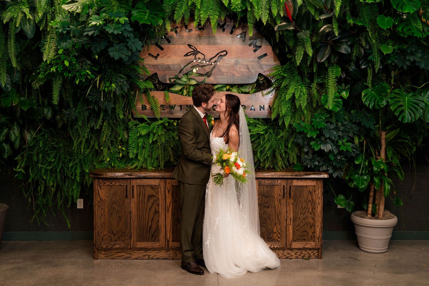 Bride and groom share romantic kiss in front of lush green living plant wall at wedding venue.