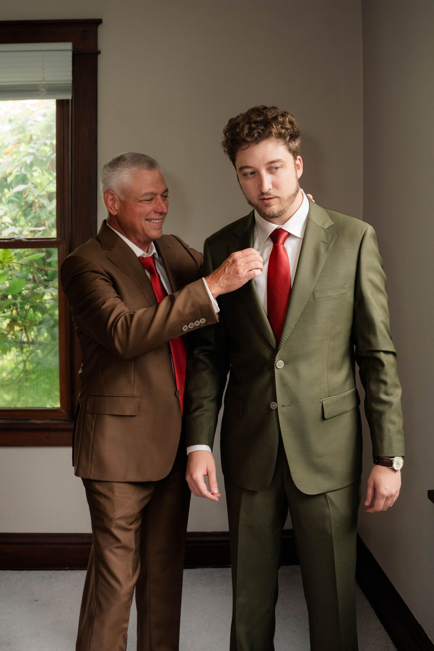 Older man in brown suit helping young man adjust his olive green suit and red tie before a formal event.