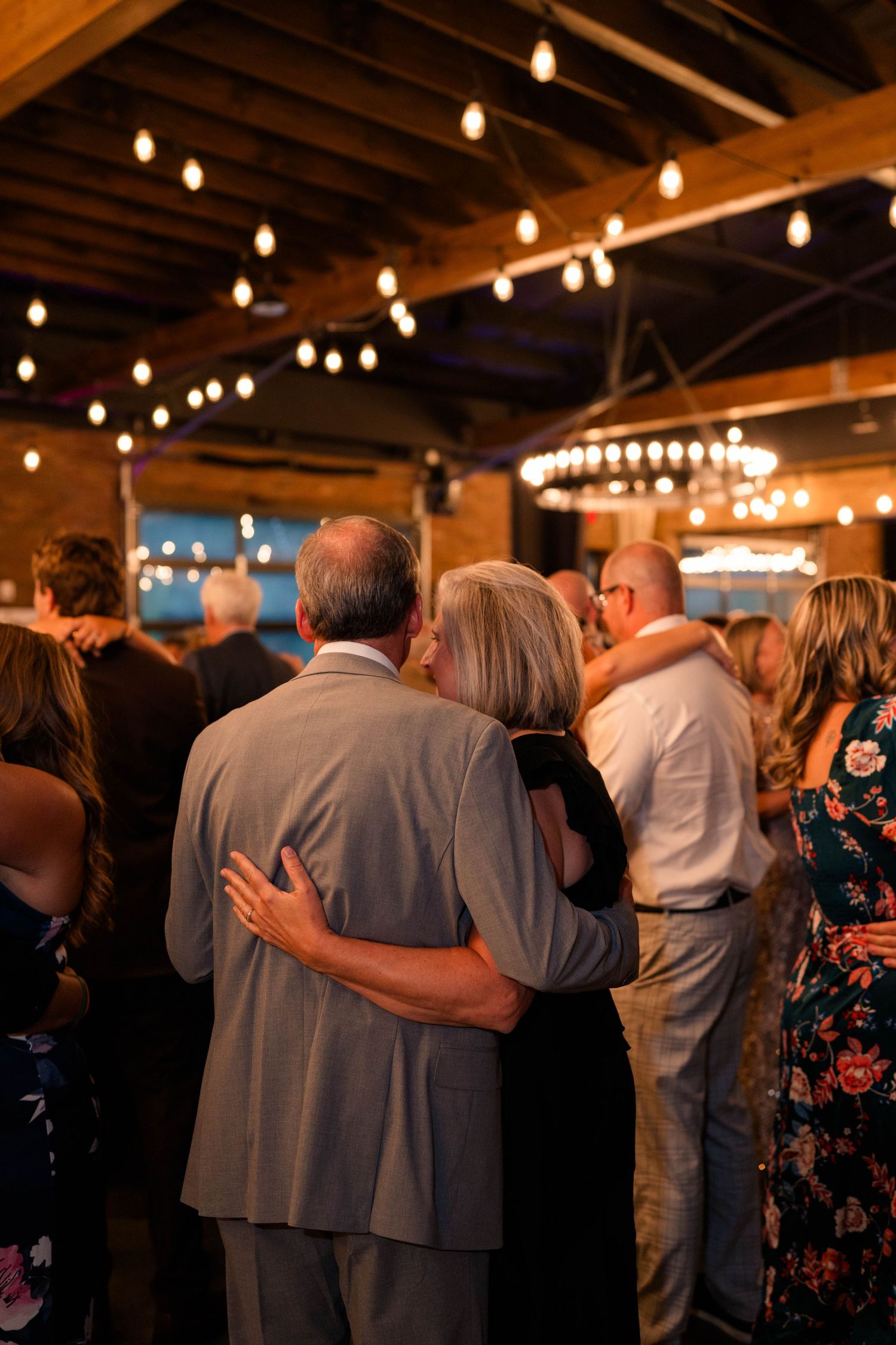 Wedding guests slow dance under vintage Edison bulb string lights in rustic reception venue.