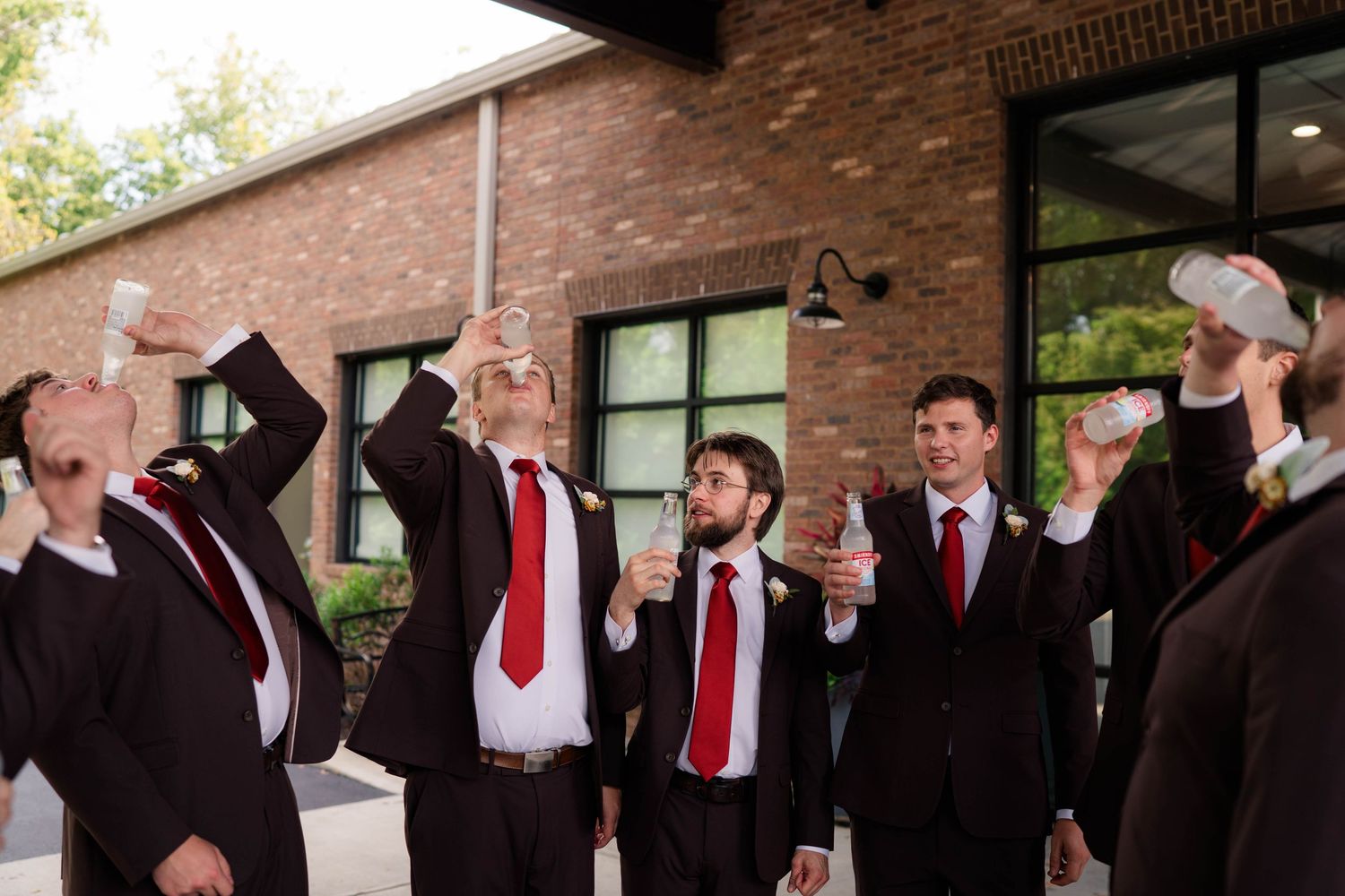 Groomsmen in dark suits and red ties raise drinks in a celebratory toast outside a brick building.