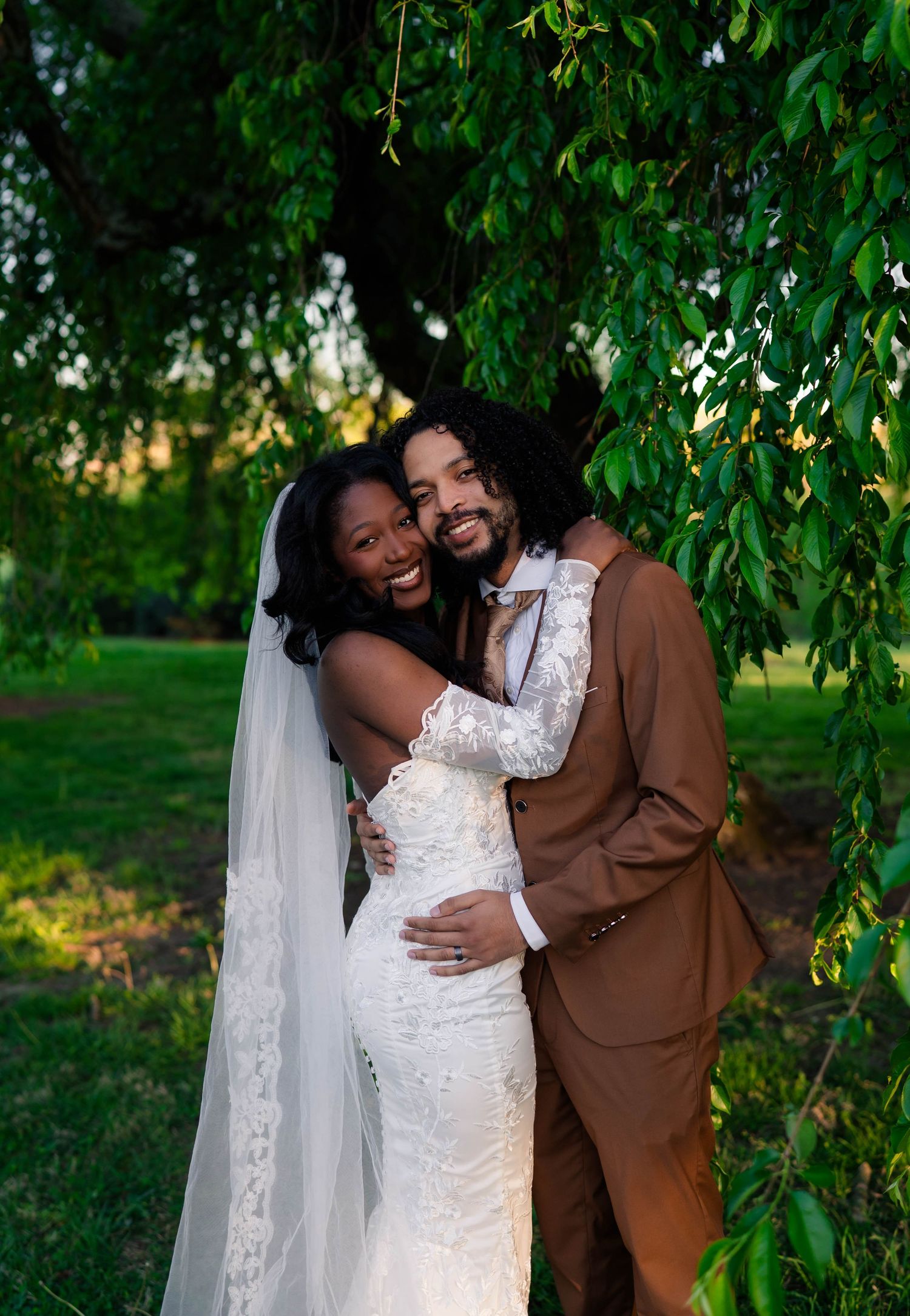 Bride in white lace gown and veil embraces groom in brown suit under weeping willow tree at outdoor wedding.