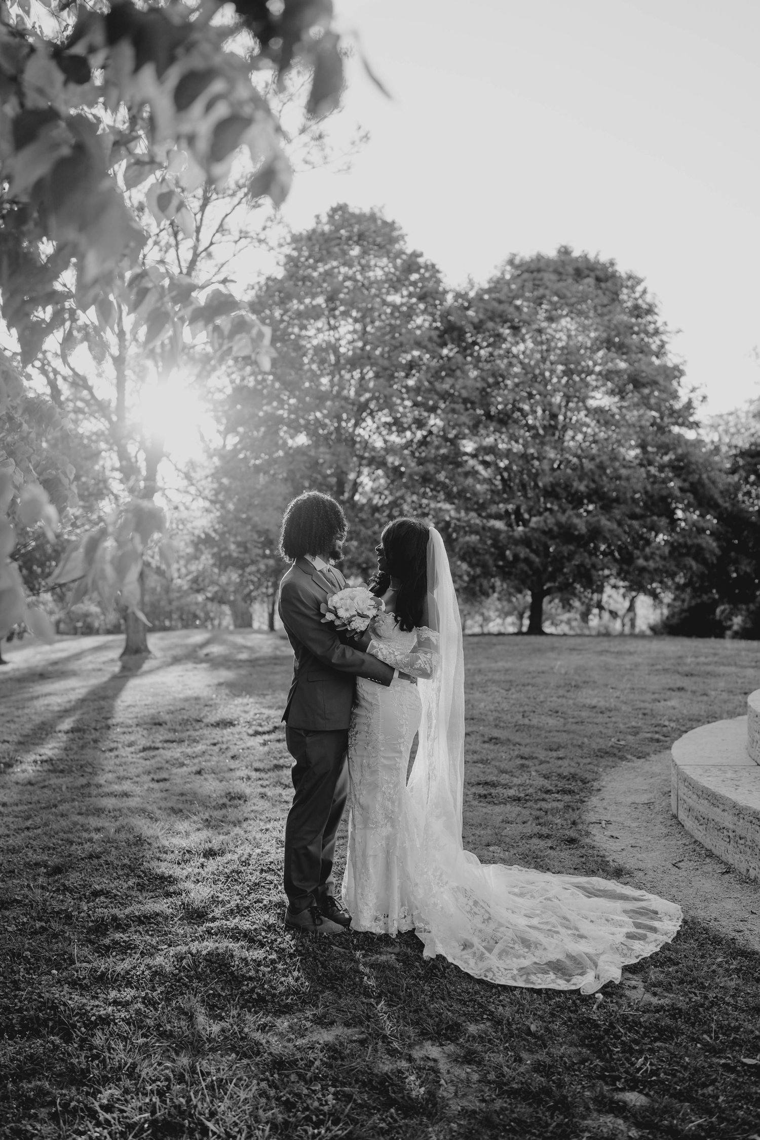 Black and white photo of a bride and groom embracing in a sunlit park, with her lace veil trailing on the grass.