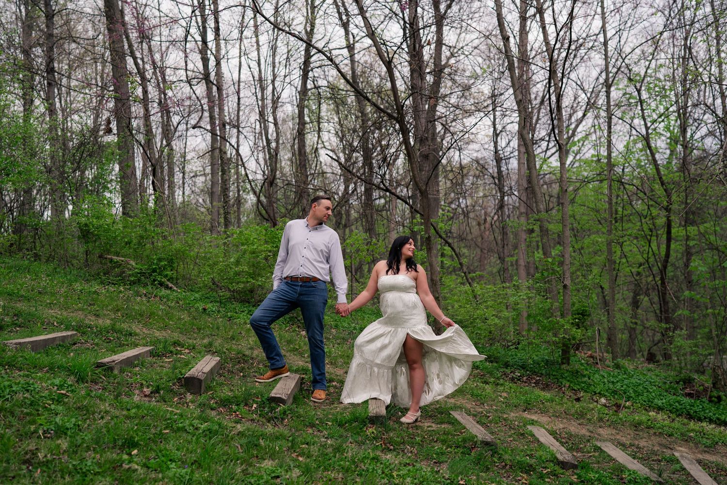 Engaged couple holding hands while walking down rustic woodland steps surrounded by spring trees.