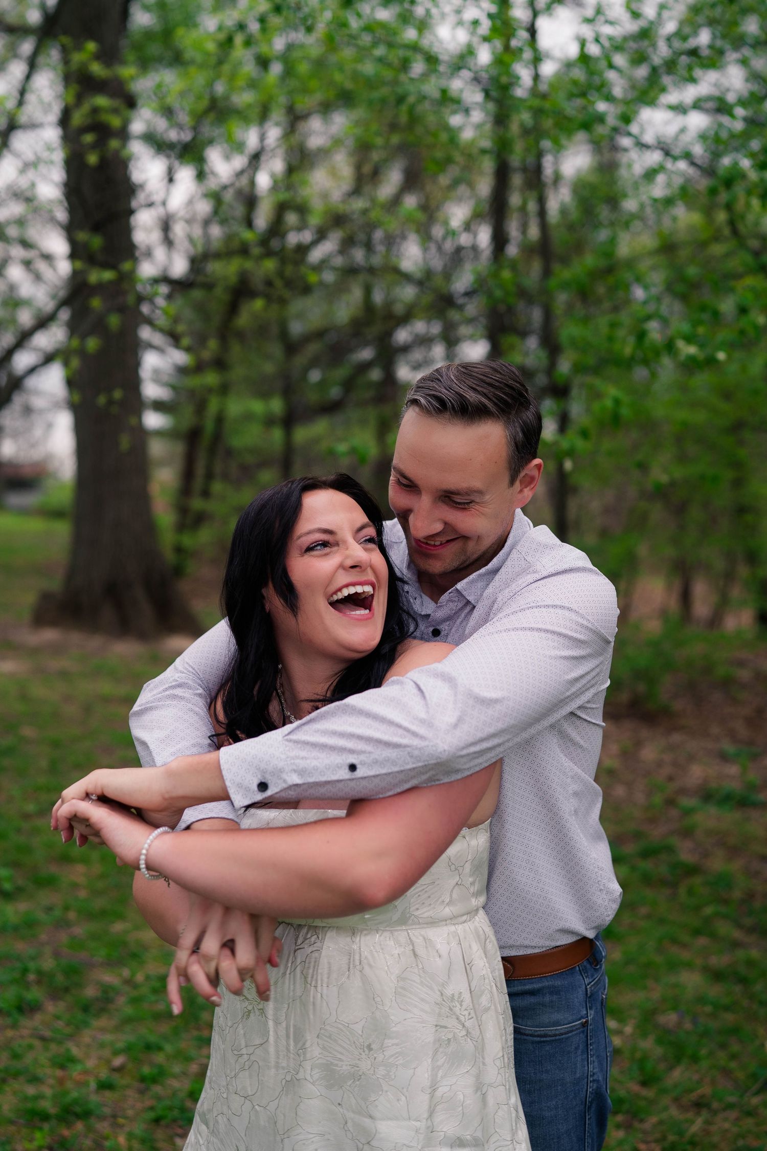 A joyful couple embraces outdoors, the woman laughing as the man hugs her from behind in a lush green park.