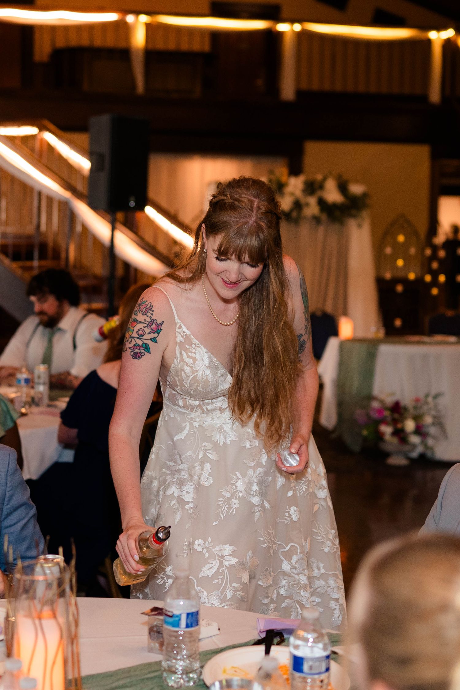 Bride in floral reception dress pouring drinks for guests at indoor wedding reception.