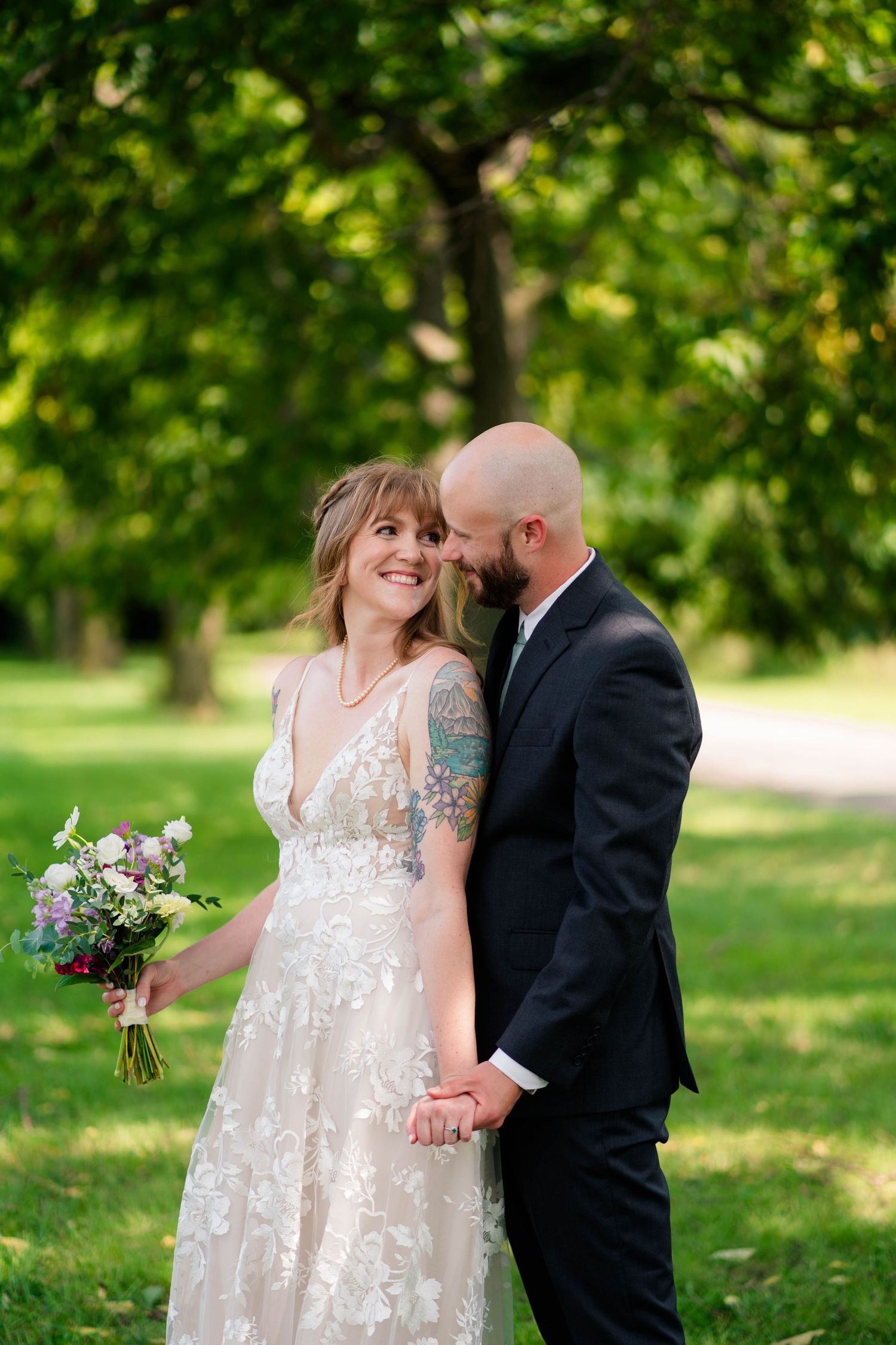Bride and groom hold hands smiling in lush green park, bride wearing floral lace gown holding bouquet.