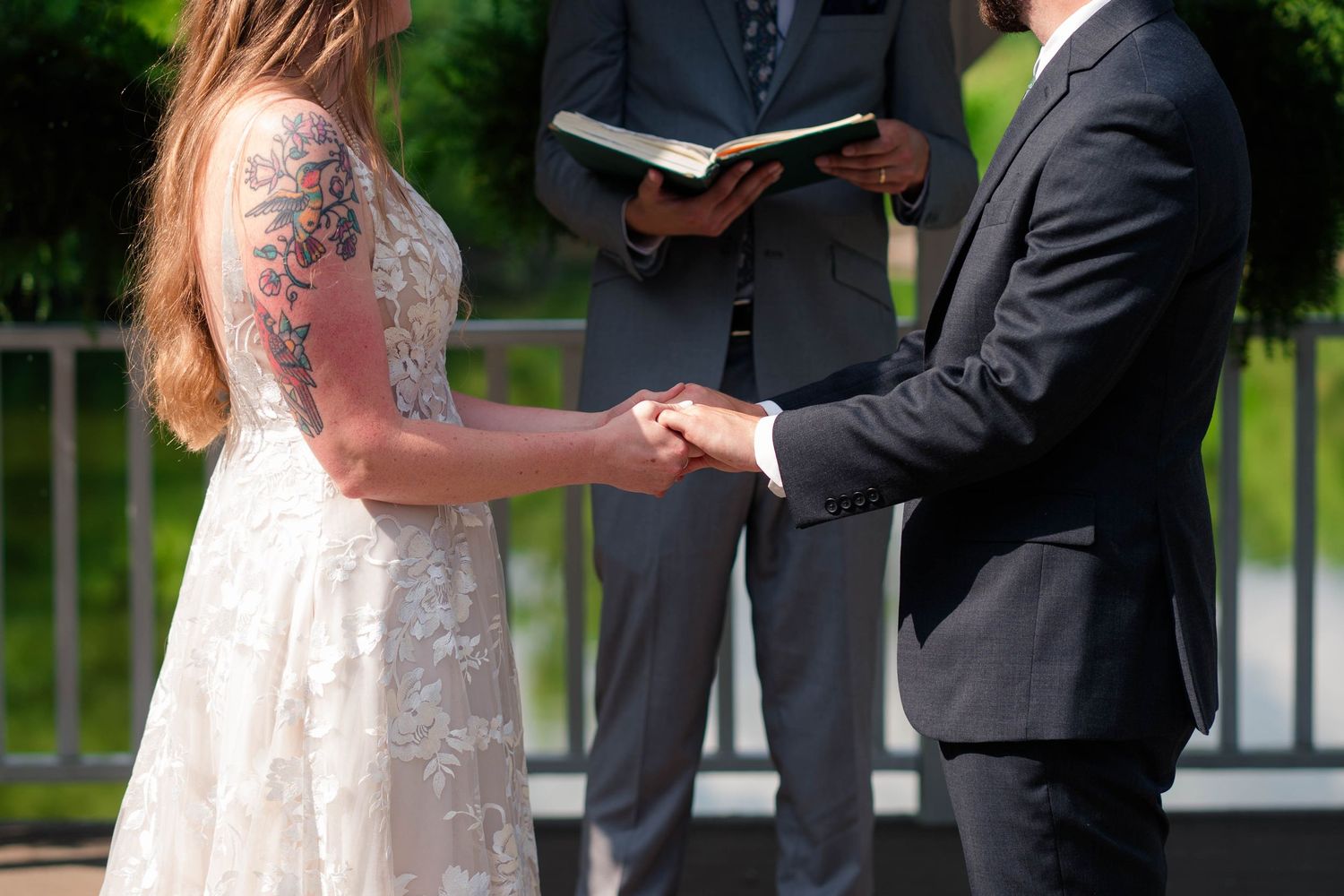 Bride with floral arm tattoo holds hands with groom while officiant reads from book during wedding ceremony.