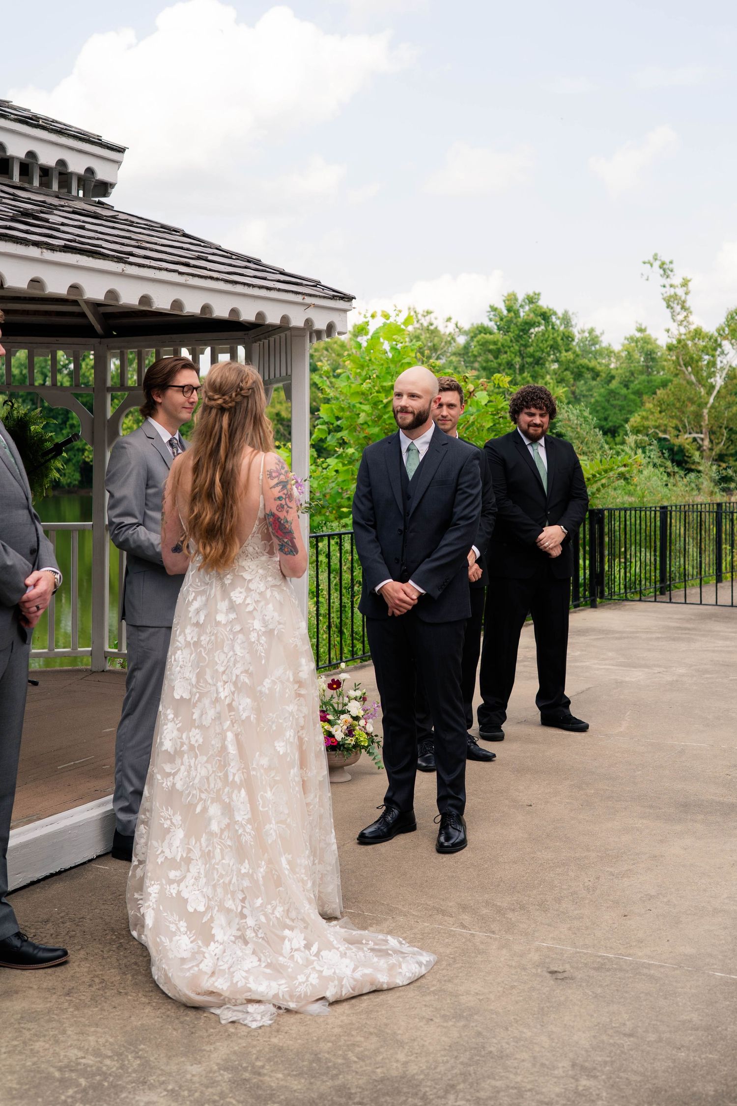 Bride in floral lace gown faces bald groom in dark suit during outdoor wedding ceremony near gazebo.