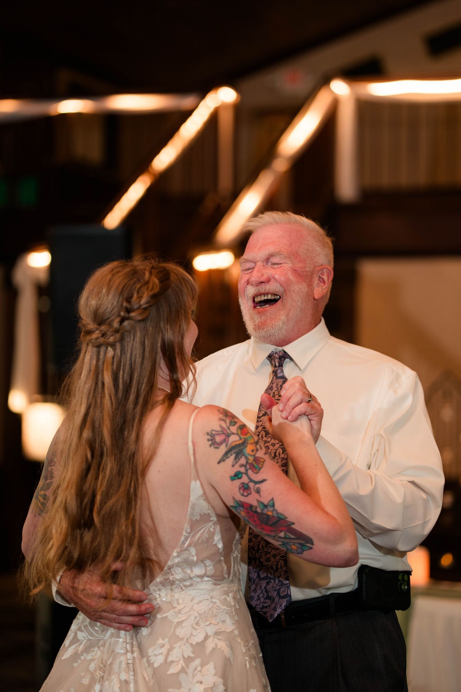 Father laughs joyfully while dancing with tattooed bride at wedding reception celebration.