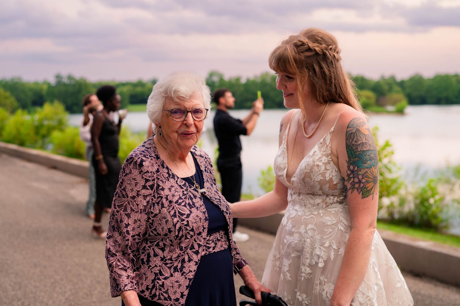 Bride with floral tattoo shares warm moment with elderly grandmother at waterfront wedding.