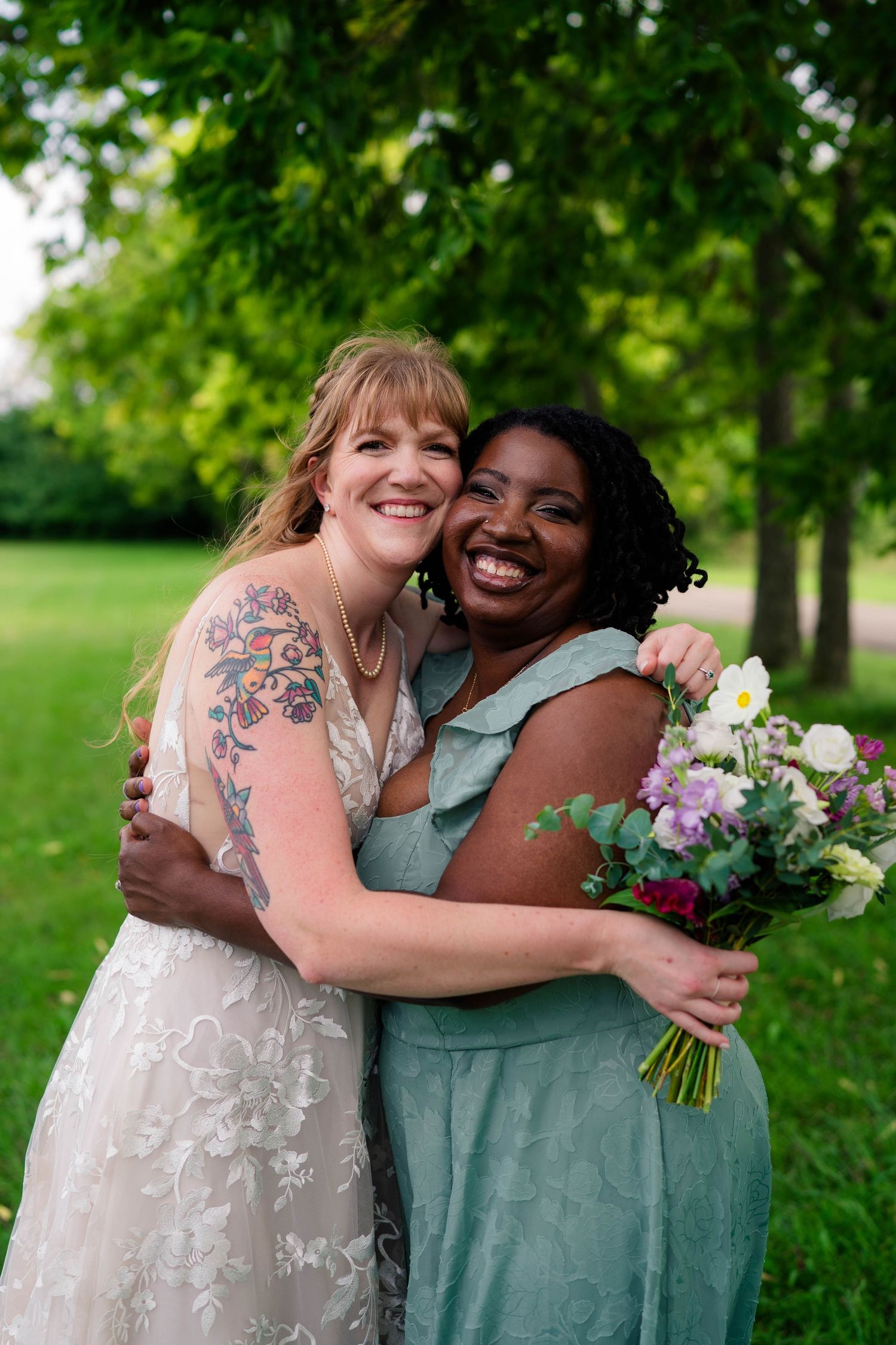 Bride with floral tattoo and bridesmaid in mint dress share a joyful hug at outdoor wedding.