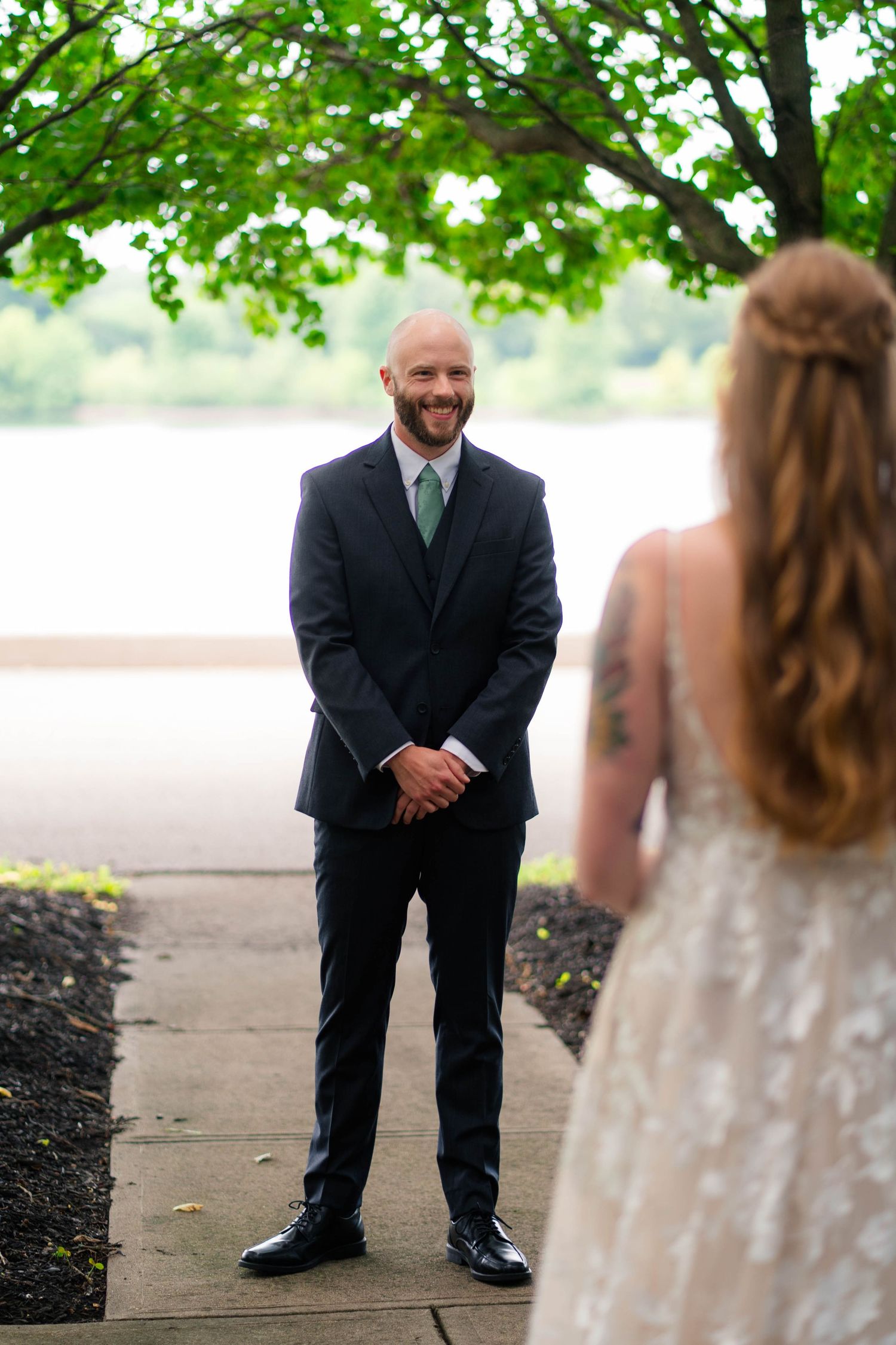 Groom in black suit smiling at bride during outdoor first look near a lake.