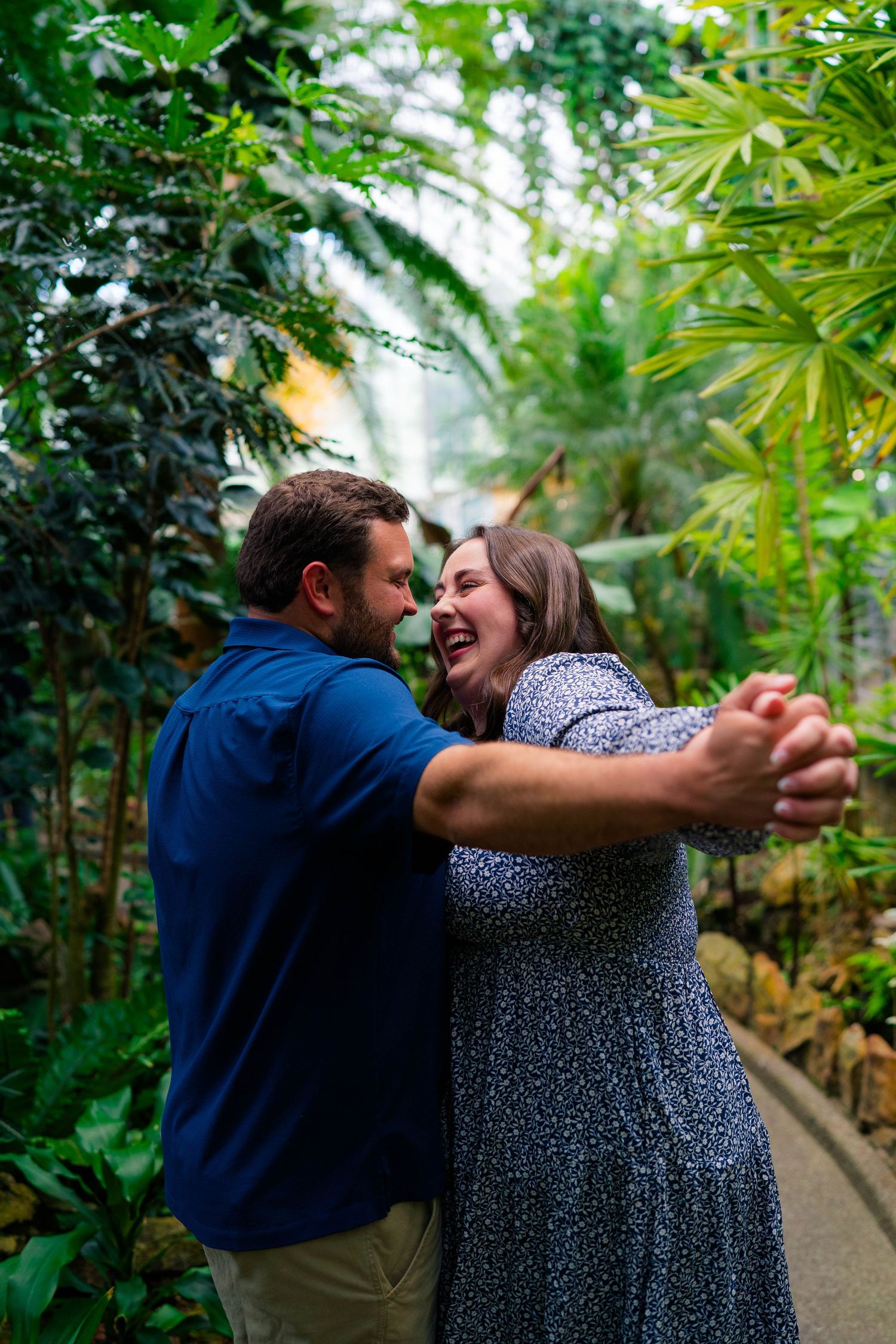 A happy couple dances and laughs together on a path surrounded by lush tropical foliage indoors.