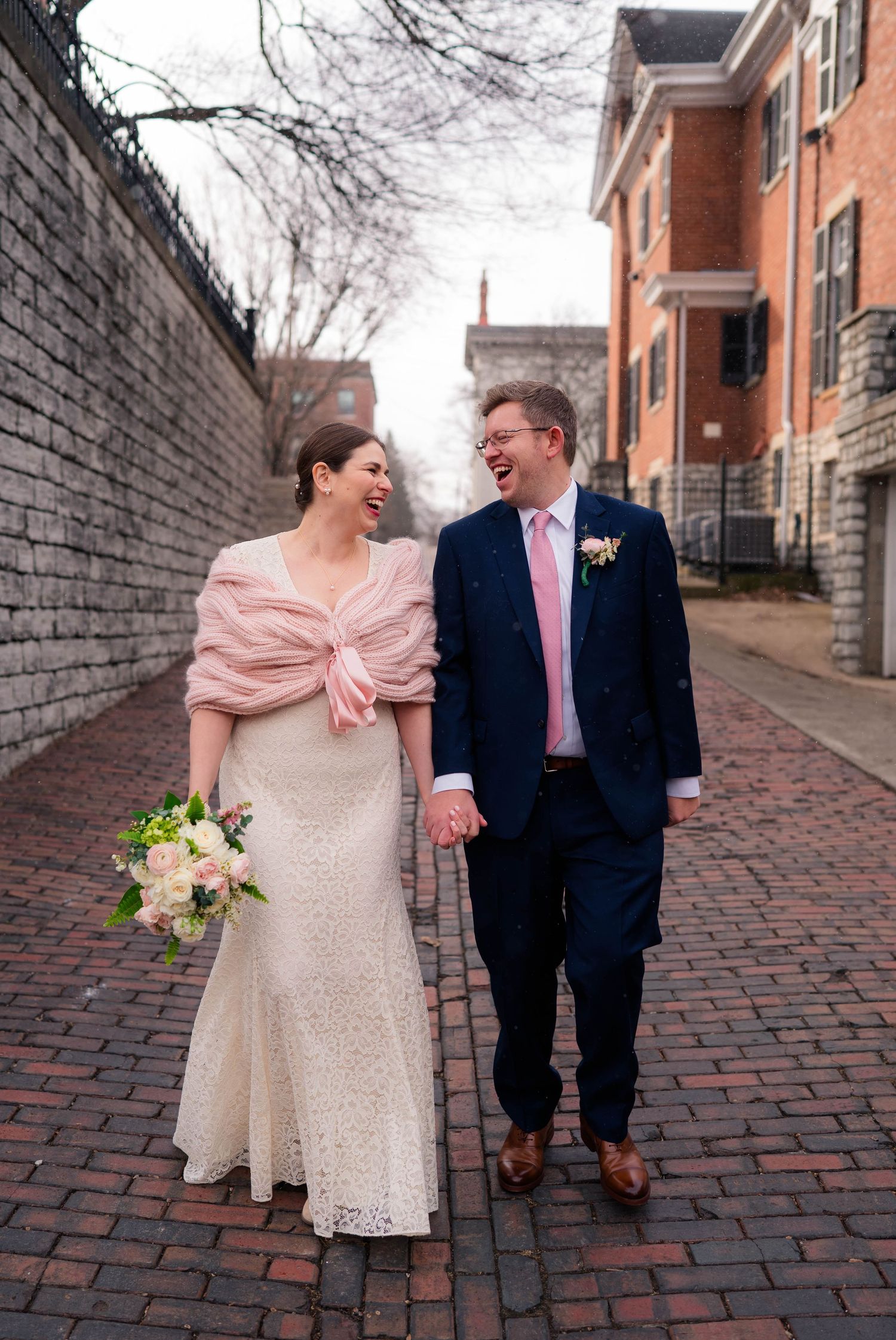 Laughing bride in lace gown with pink shawl and groom in navy suit share a joyful moment on a brick path.
