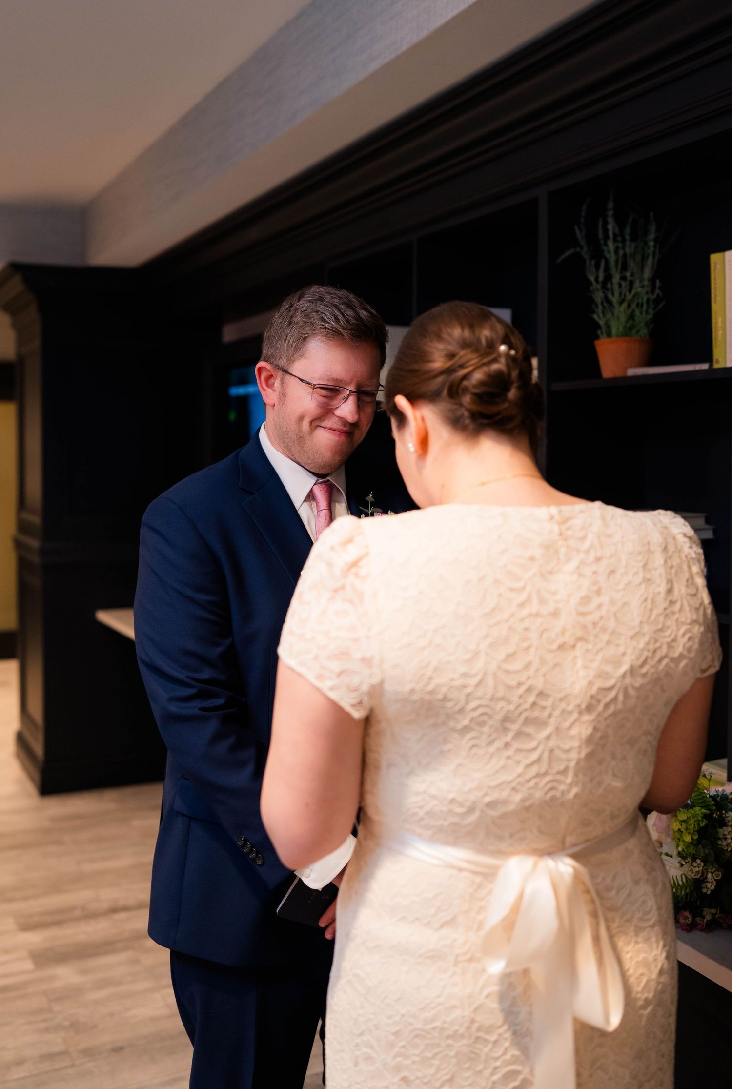 Smiling groom in navy suit holding brides hands tenderly during emotional private vow exchange.