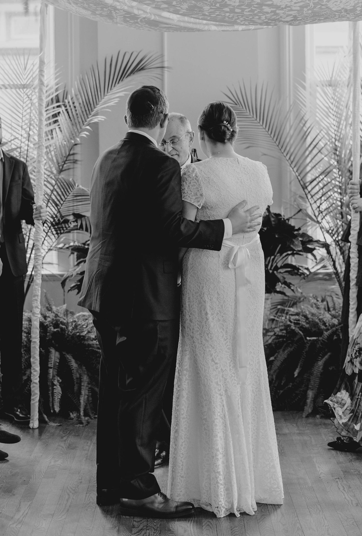 Black and white photo of officiant embracing bride and groom from behind under chuppah after wedding ceremony.