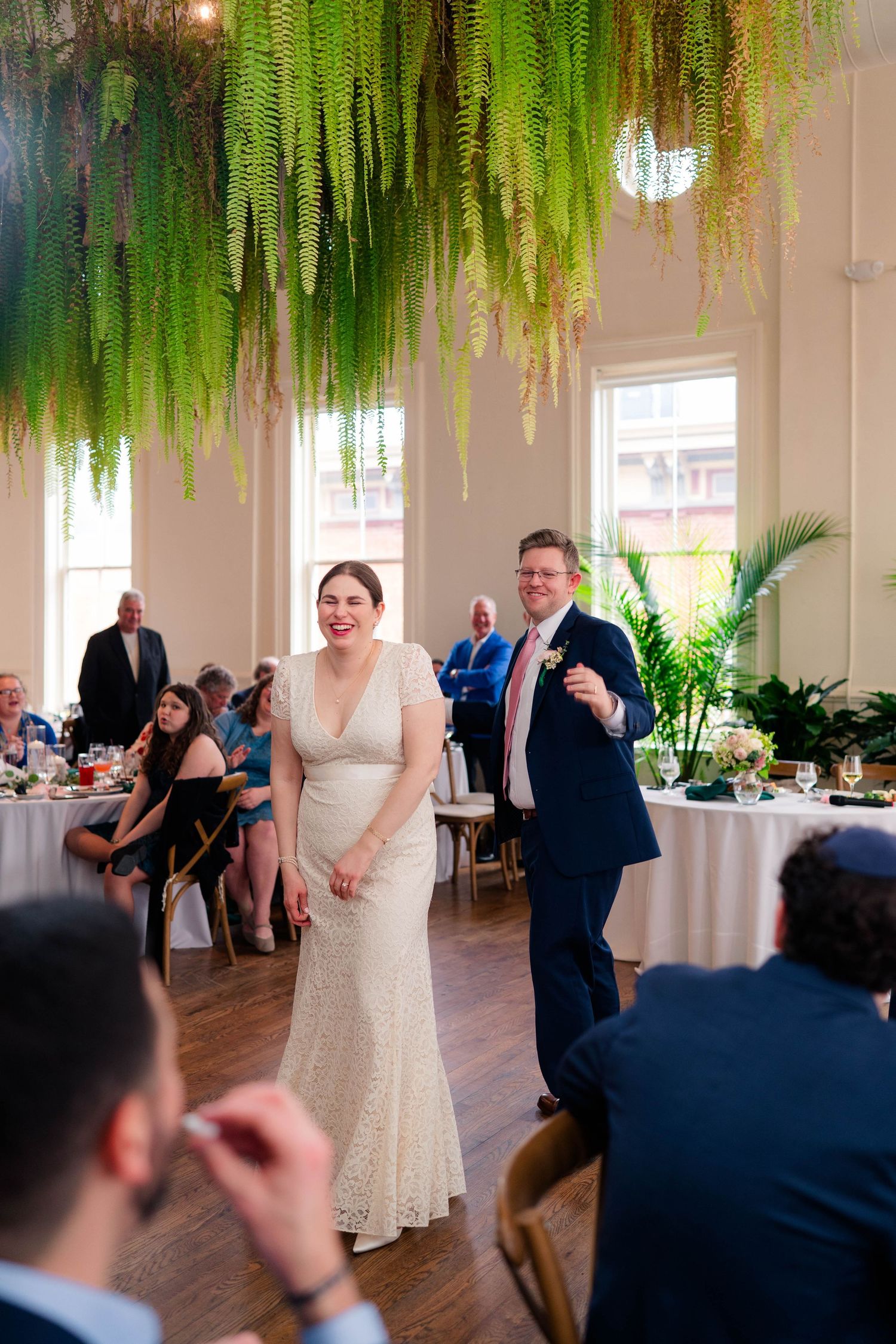 Bride and groom enter wedding reception hall decorated with hanging fern greenery installation above dance floor.