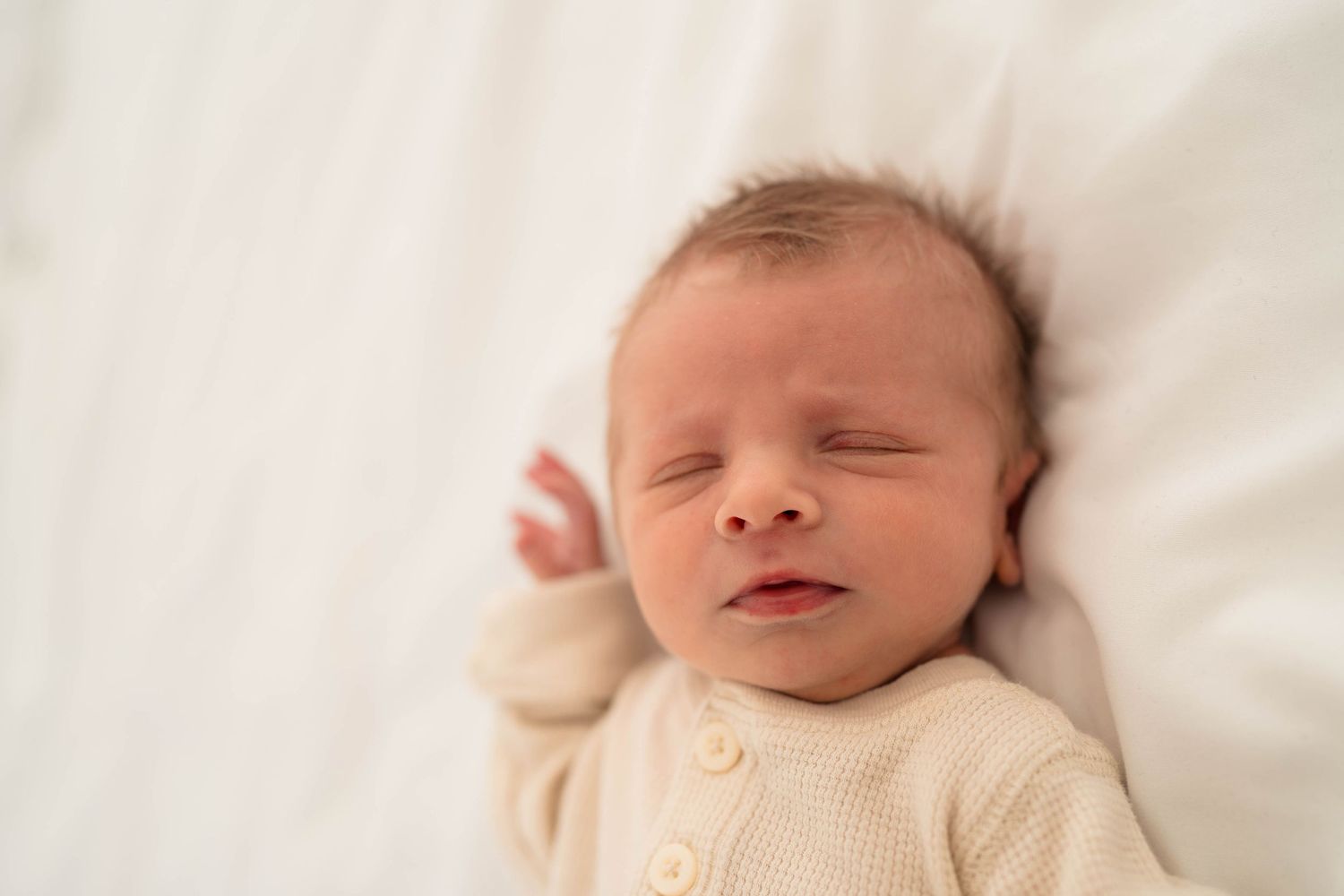 Peaceful newborn baby in cream onesie sleeping on white blanket with soft natural lighting.