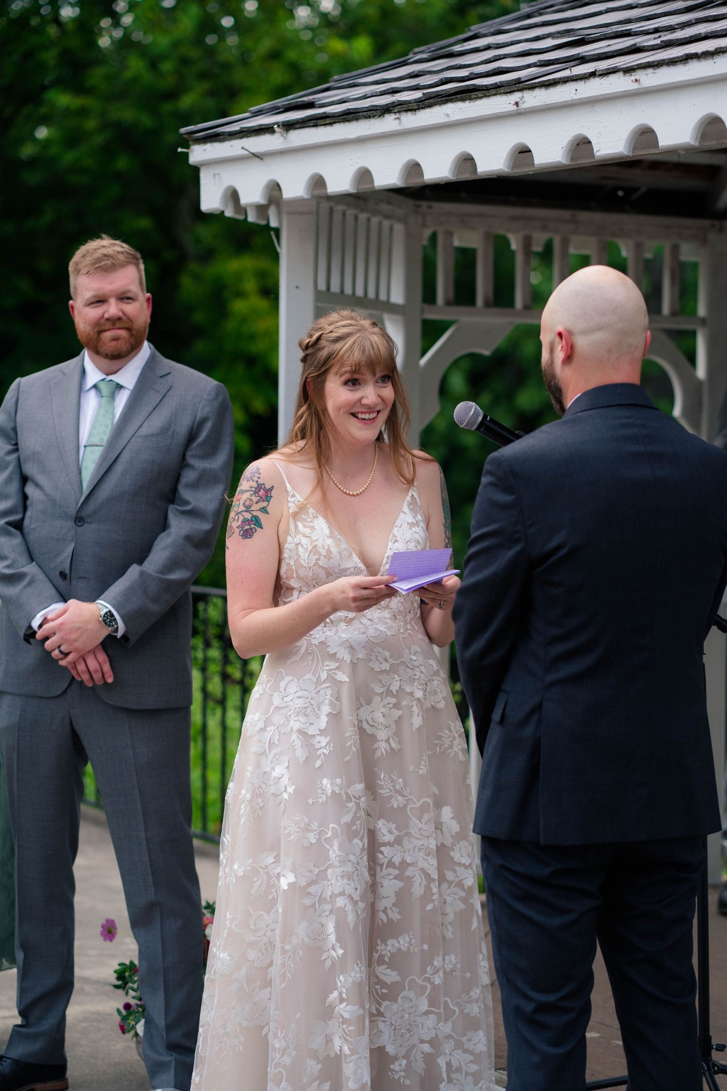 Bride smiling while reading vows at outdoor gazebo wedding ceremony with groom and officiant.