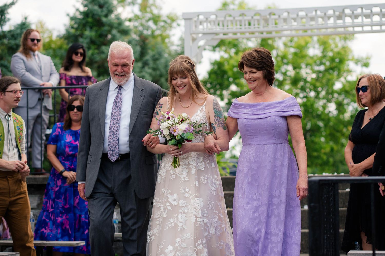 Bride walks down the aisle with her parents at an outdoor wedding ceremony.