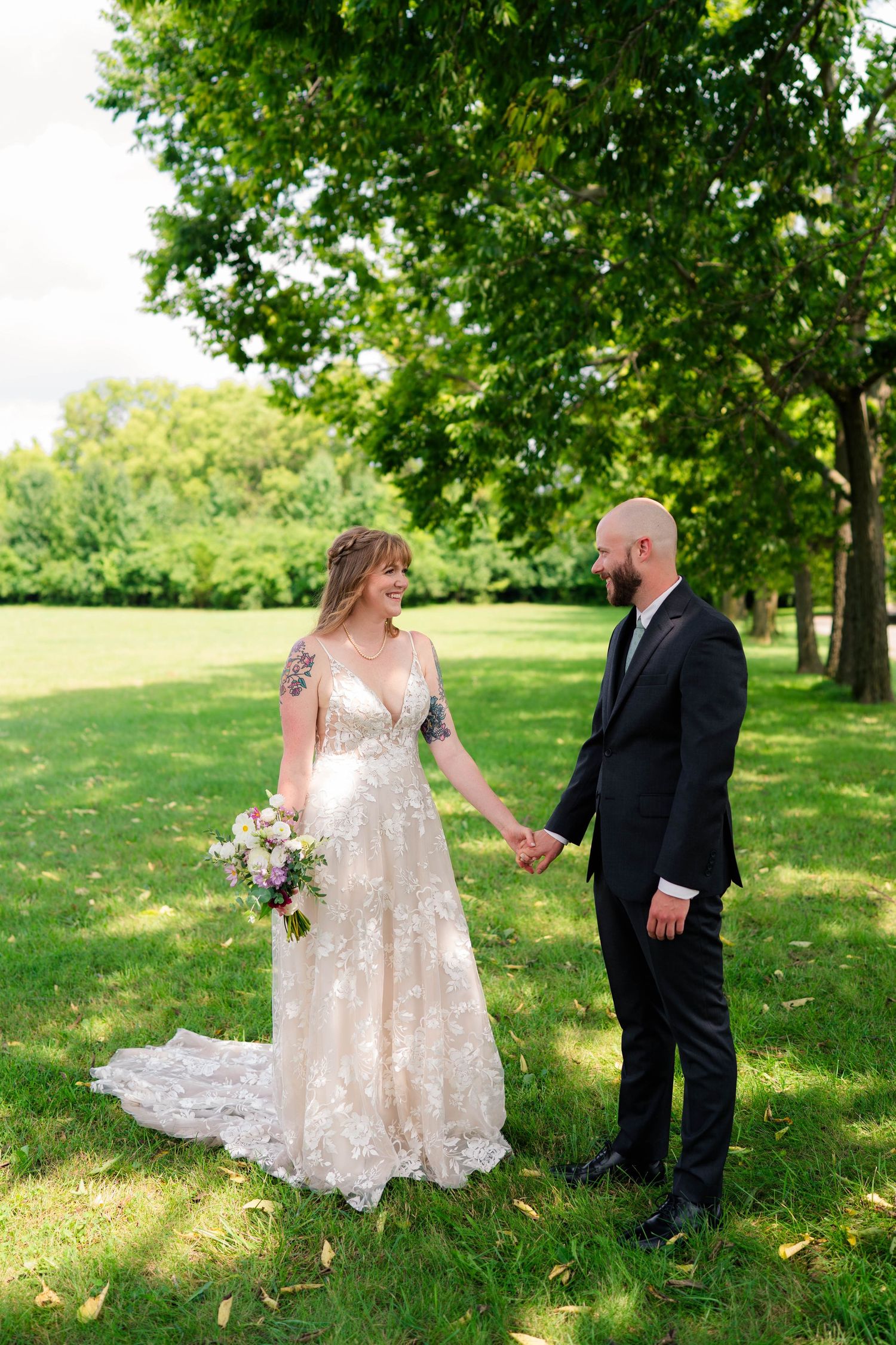 Bride in floral lace gown with arm tattoo holds bouquet while holding hands with bald groom on green lawn.
