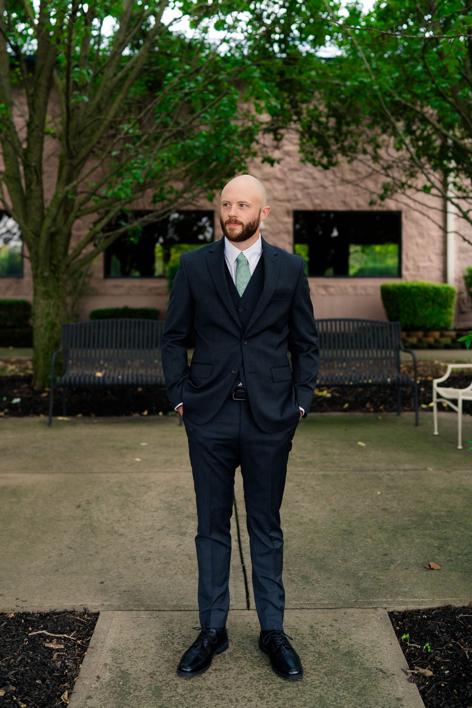 Bald groom in navy suit with sage green tie stands confidently outdoors before wedding ceremony.