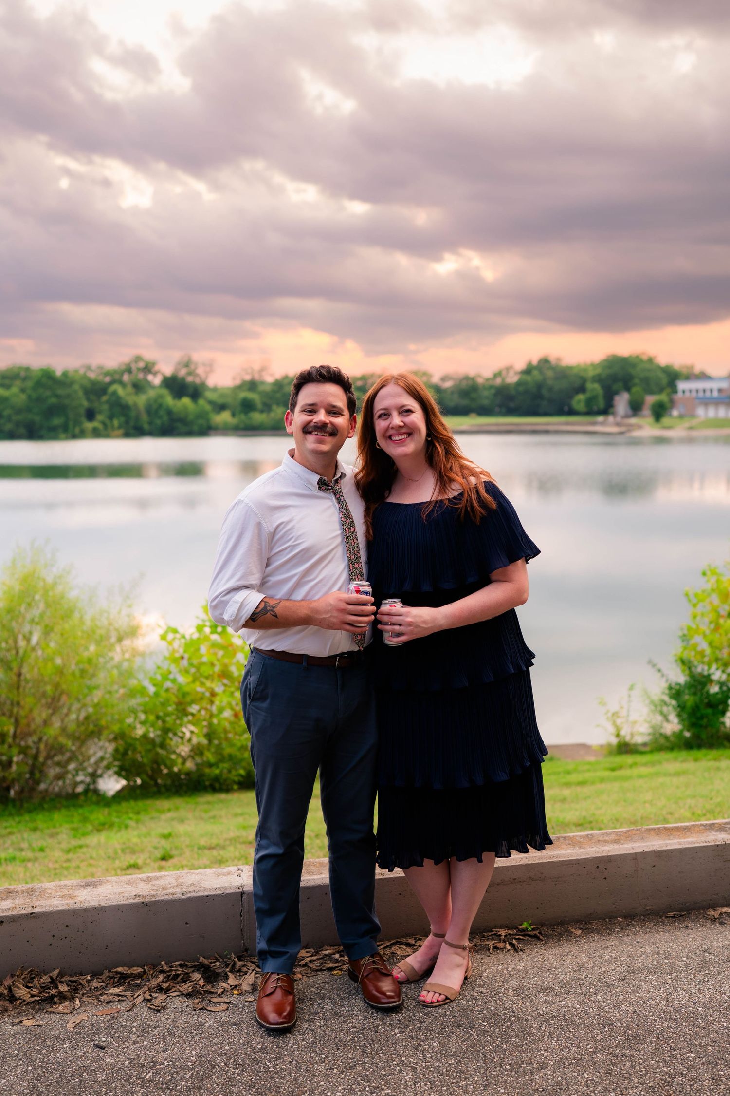 Smiling couple poses together at sunset by a lake with dramatic pink and purple clouds.