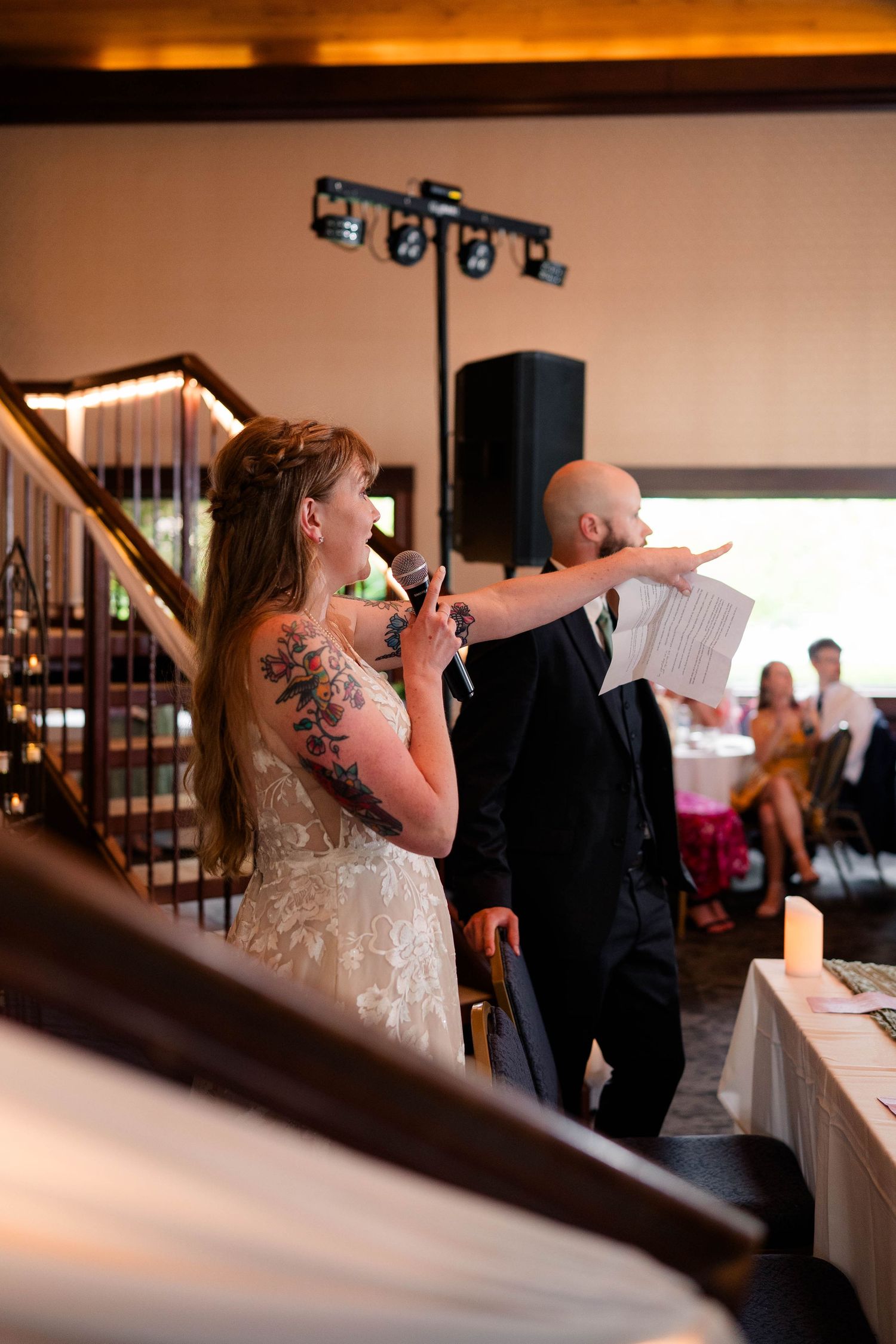 Tattooed bride speaks into microphone beside bald groom pointing during wedding reception speech.