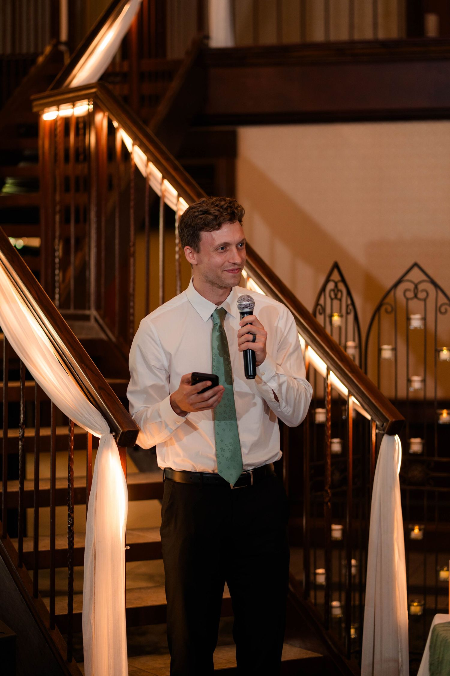 Groomsman in white shirt and green tie gives toast holding microphone at wedding reception.