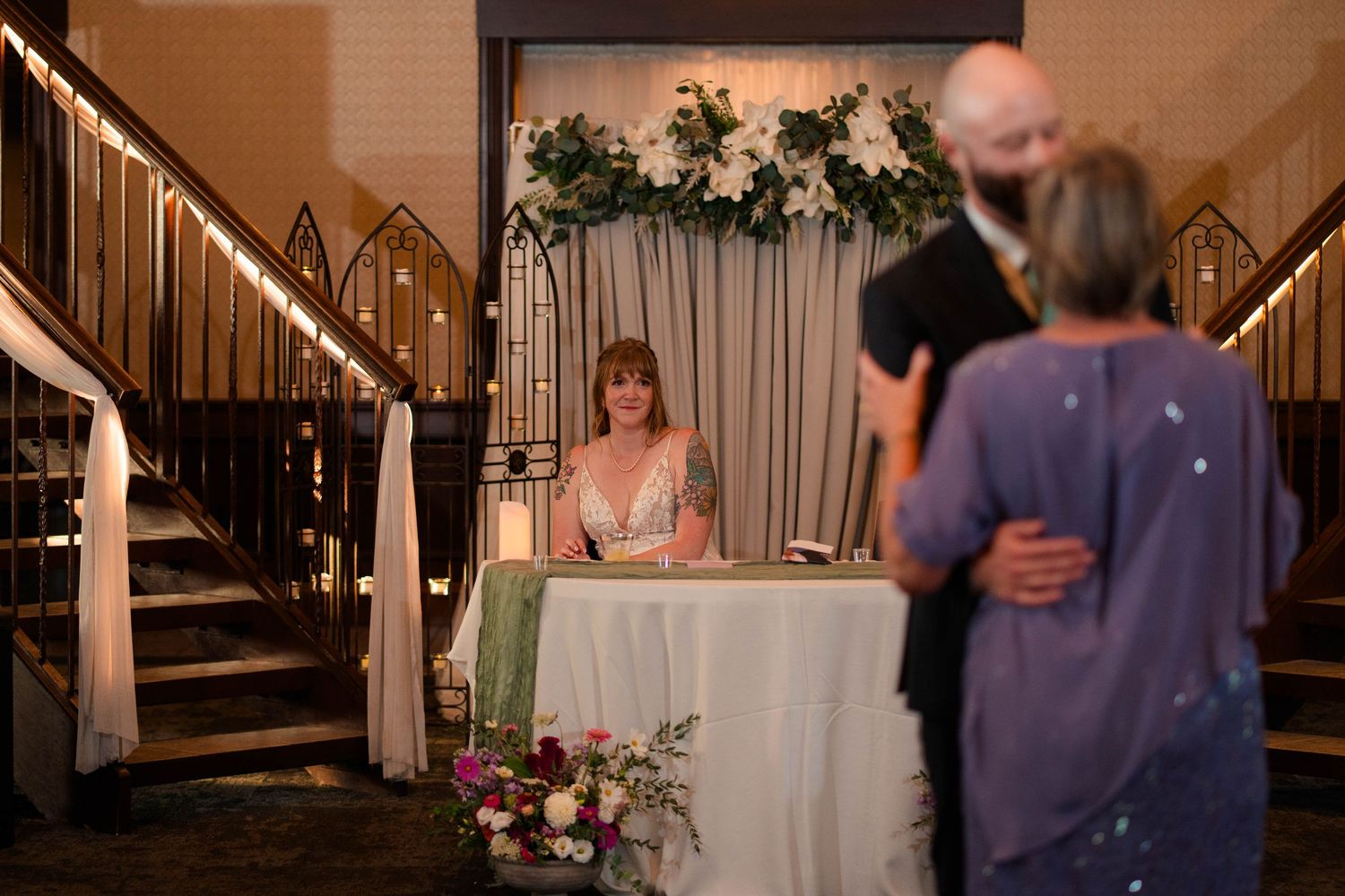 Bride sits at sweetheart table as couple dances in foreground at romantic indoor wedding reception.