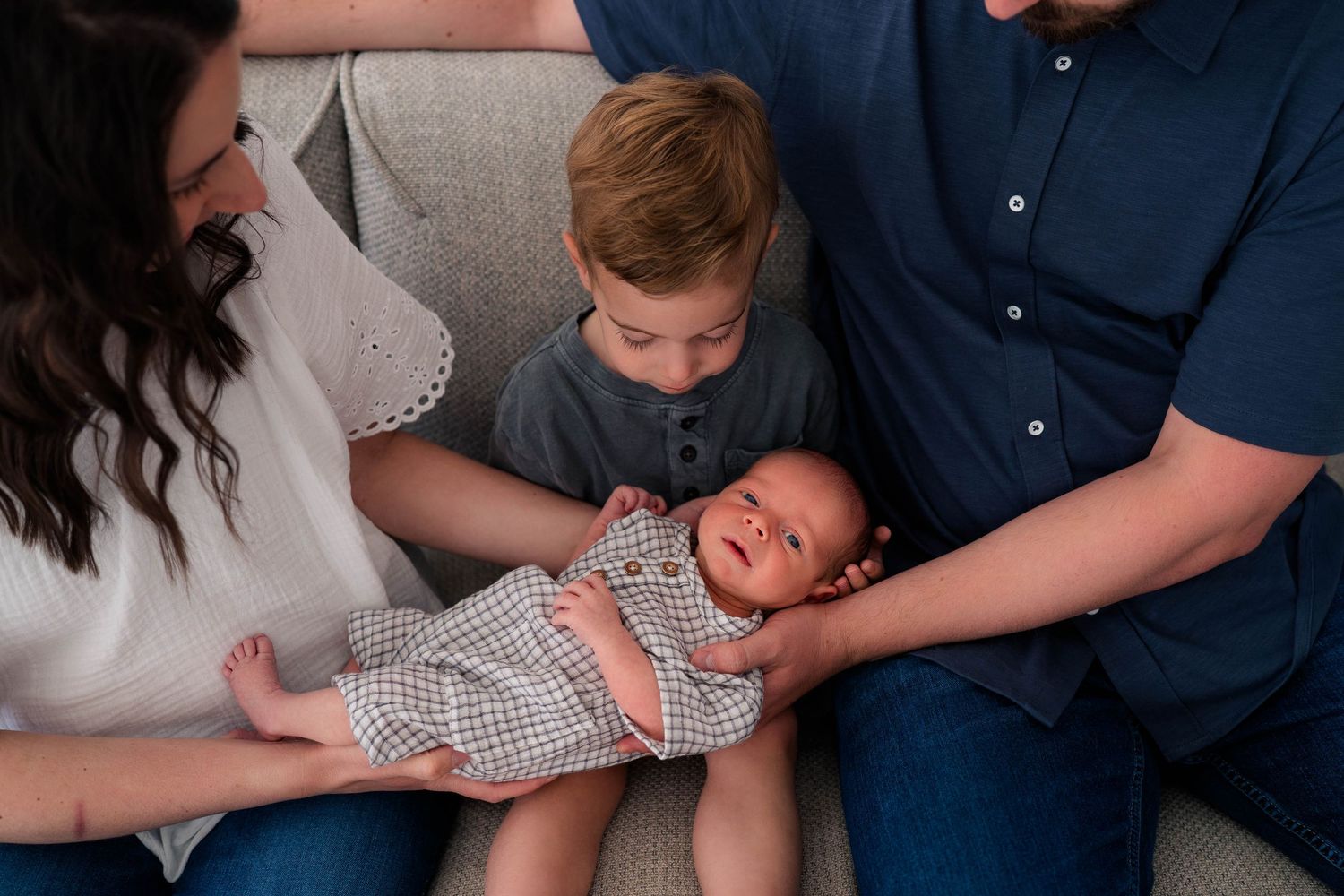 Family of four with parents, toddler boy, and newborn baby sitting together on a couch for a studio portrait session.