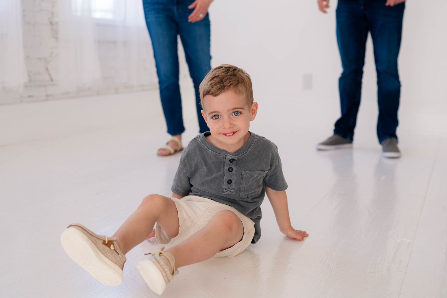 A cheerful toddler boy sits on a white floor smiling, with adults standing behind him.