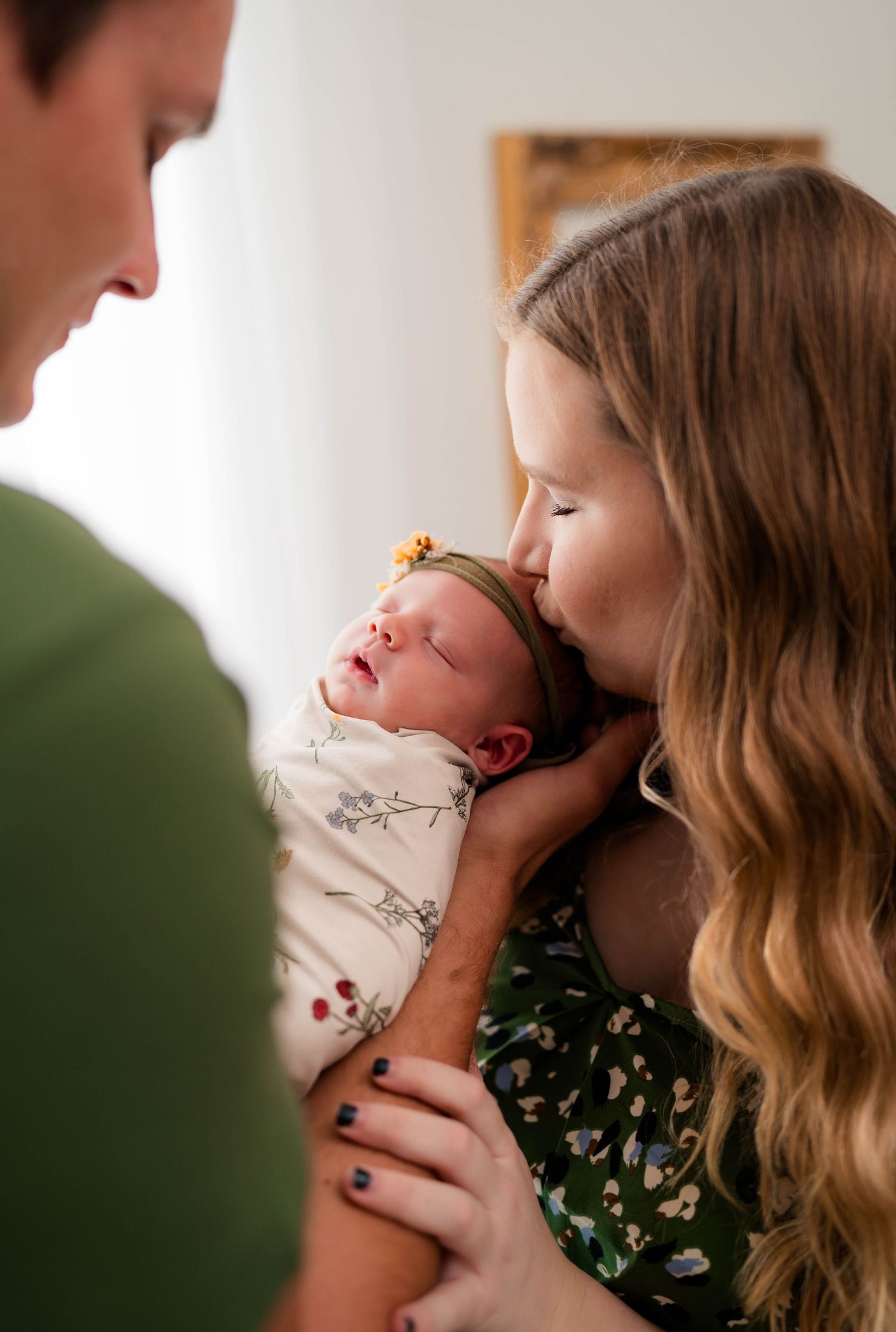 Parents lovingly hold their swaddled newborn as mother kisses baby's head in warm indoor setting.
