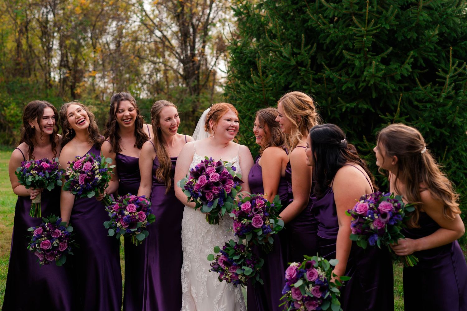 Bride laughs with bridesmaids in deep purple gowns holding lush purple and pink bouquets outdoors.