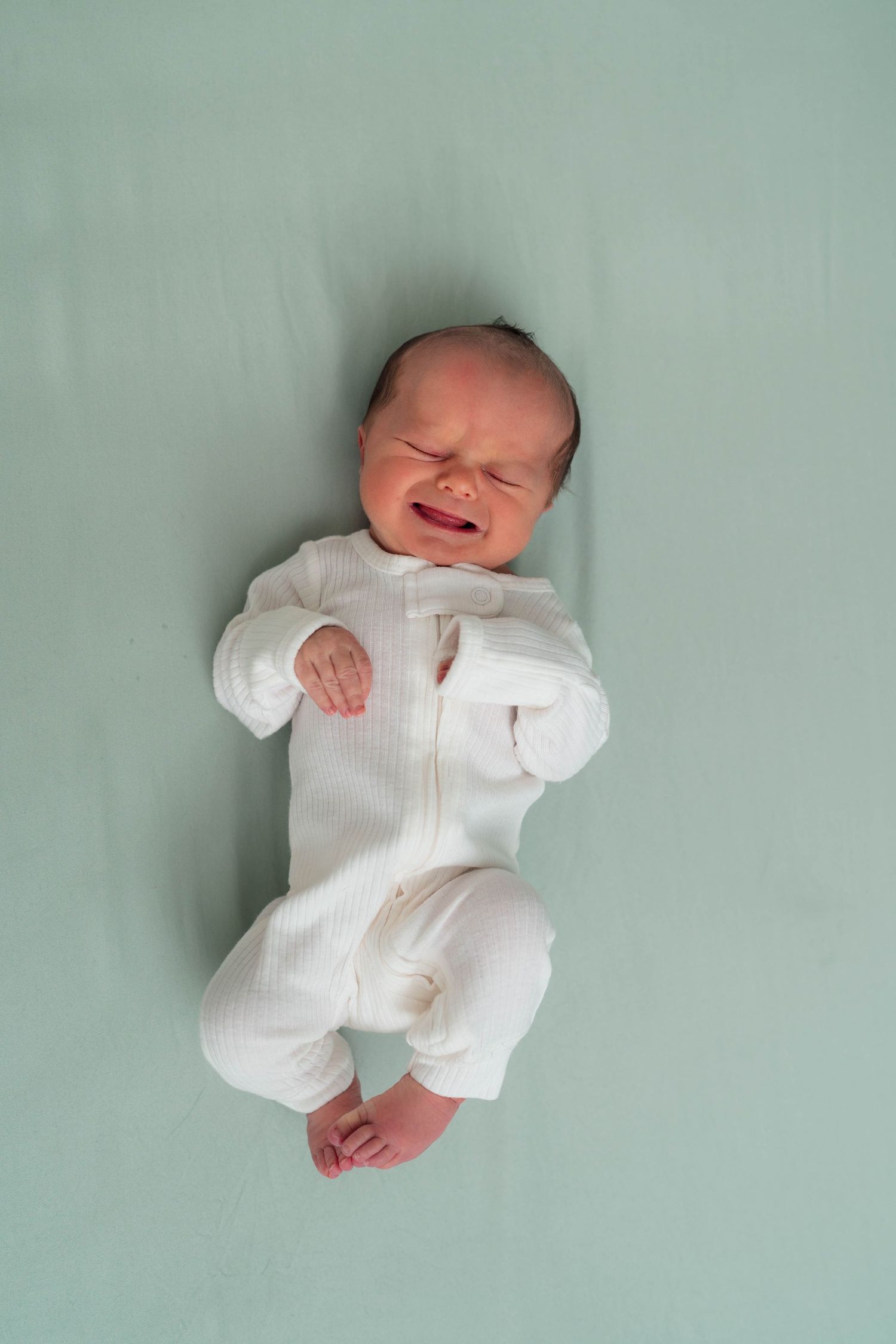 Crying newborn baby lying on teal blanket wearing white ribbed zip-up onesie.