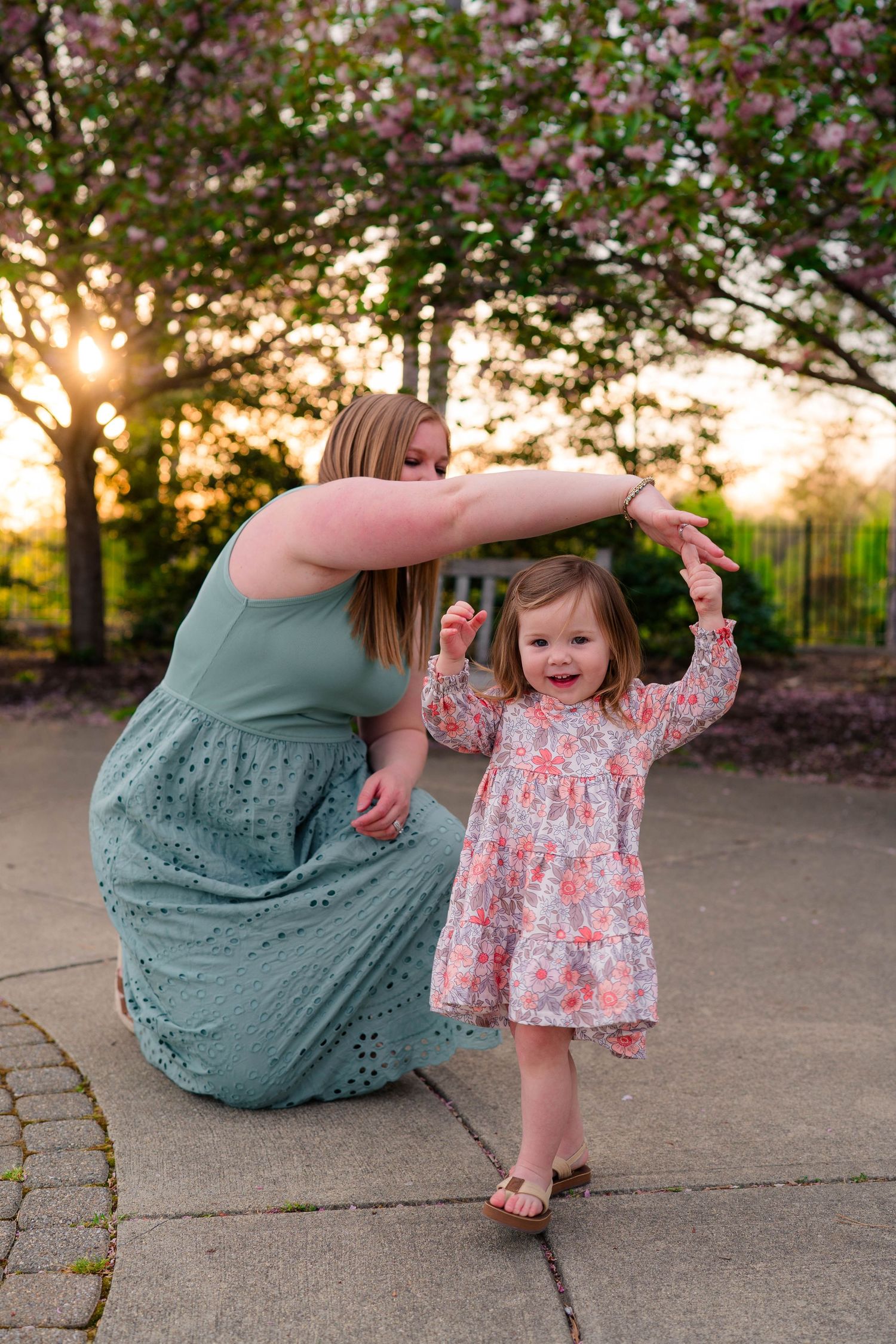 Mother helps toddler girl dance at sunset near blooming pink trees in a garden setting.