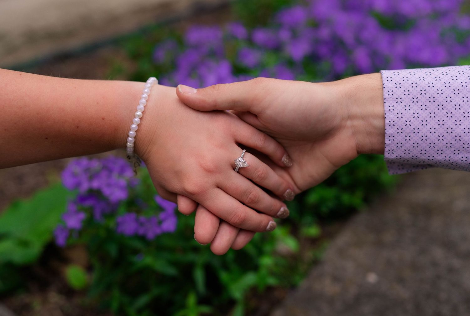 Engaged couple holding hands showing diamond engagement ring and pearl bracelet near purple flowers.