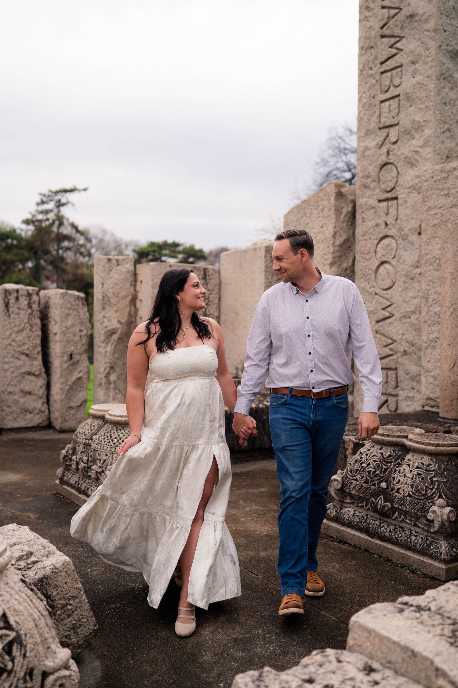 Couple holding hands and walking among stone ruins, woman in white dress and man in blue jeans, smiling at each other.