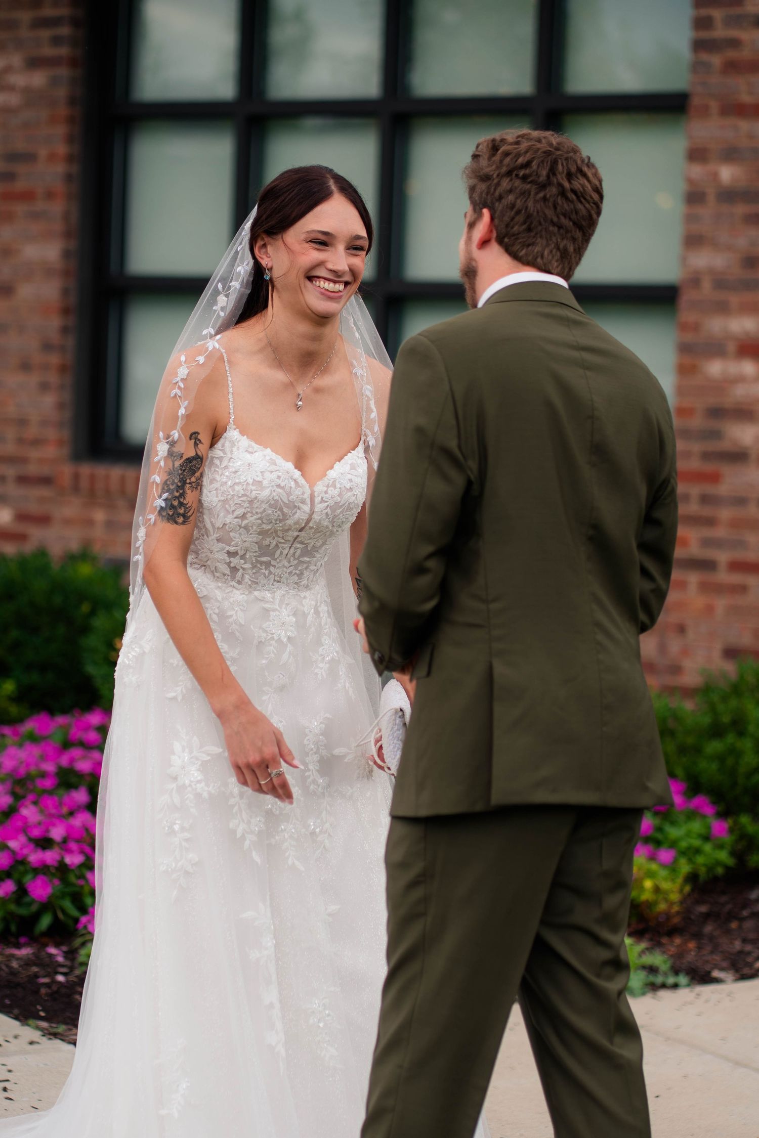 Radiant bride in lace wedding dress laughing during first look with groom in olive green suit.