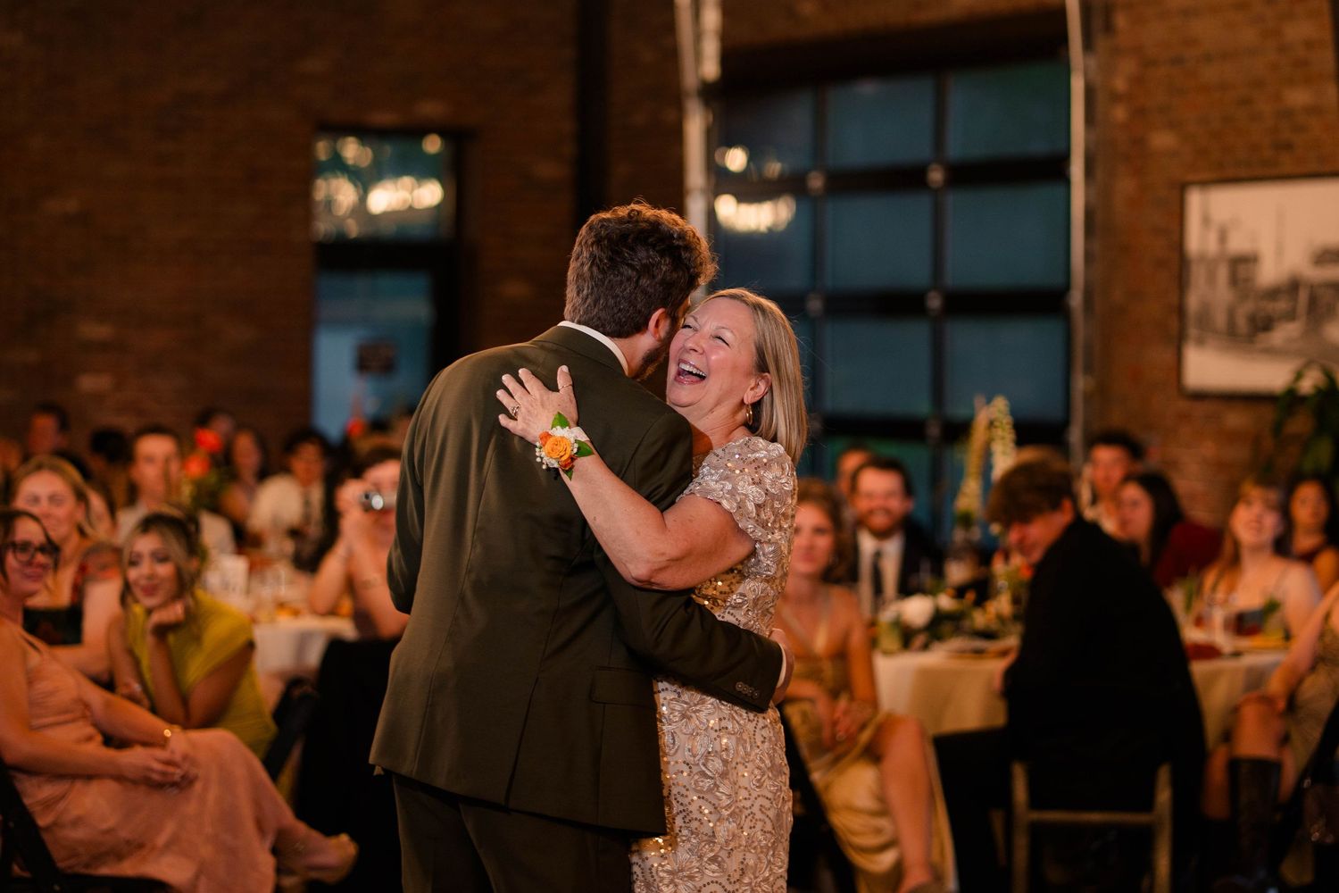 Groom dancing with his mother under warm lights surrounded by wedding reception guests.